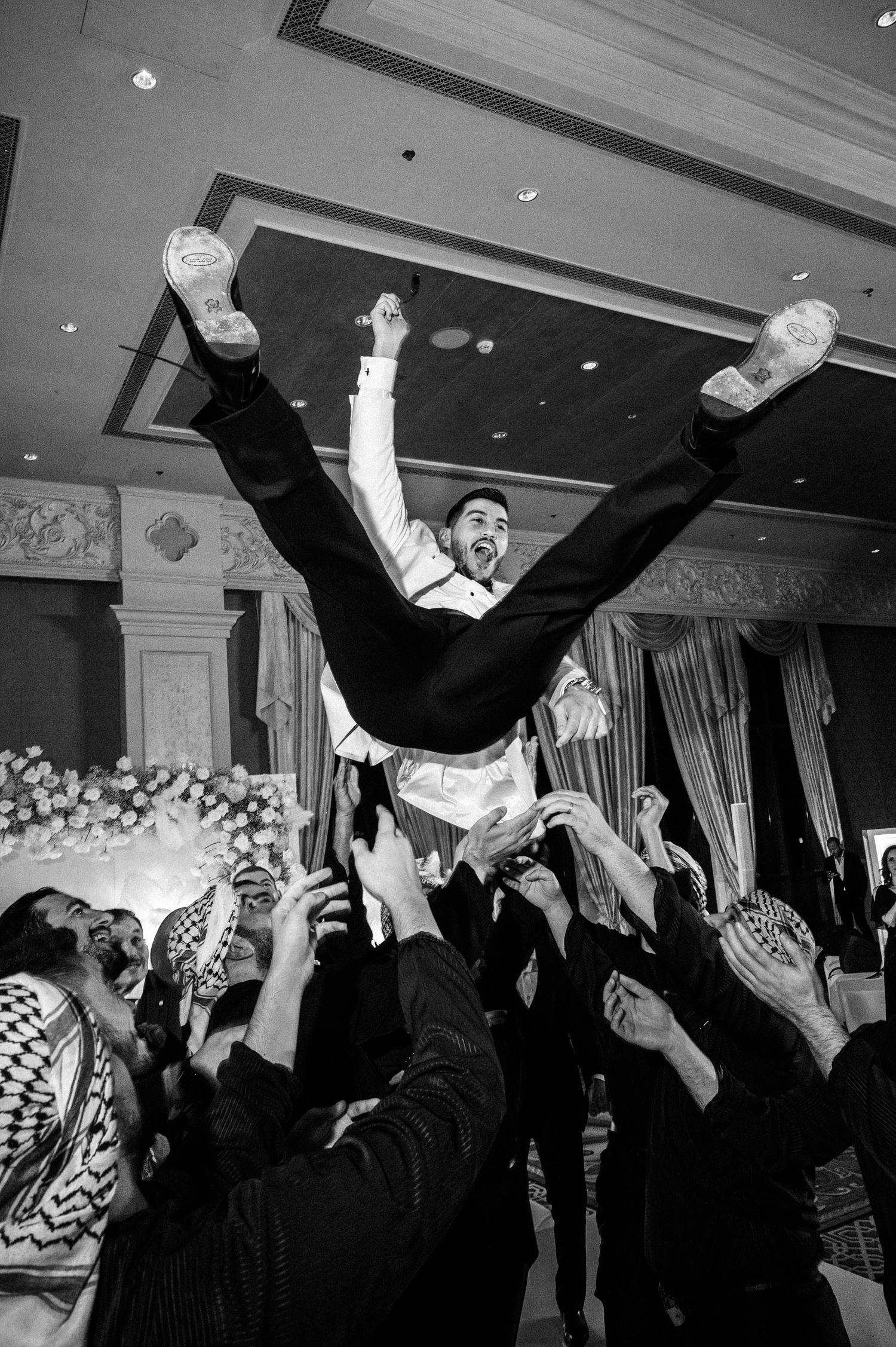 Wedding guests toss and throw the groom in the air during a joyful celebration at an indoor reception venue.