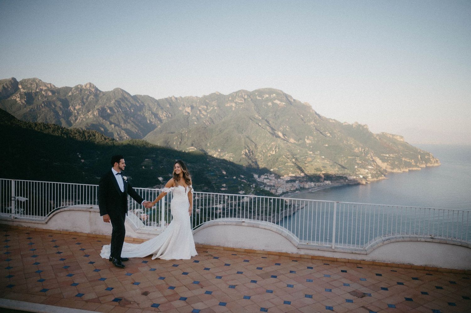 A wedding couple stands on a terrace overlooking the stunning Amalfi Coast mountains and Mediterranean Sea in Italy.