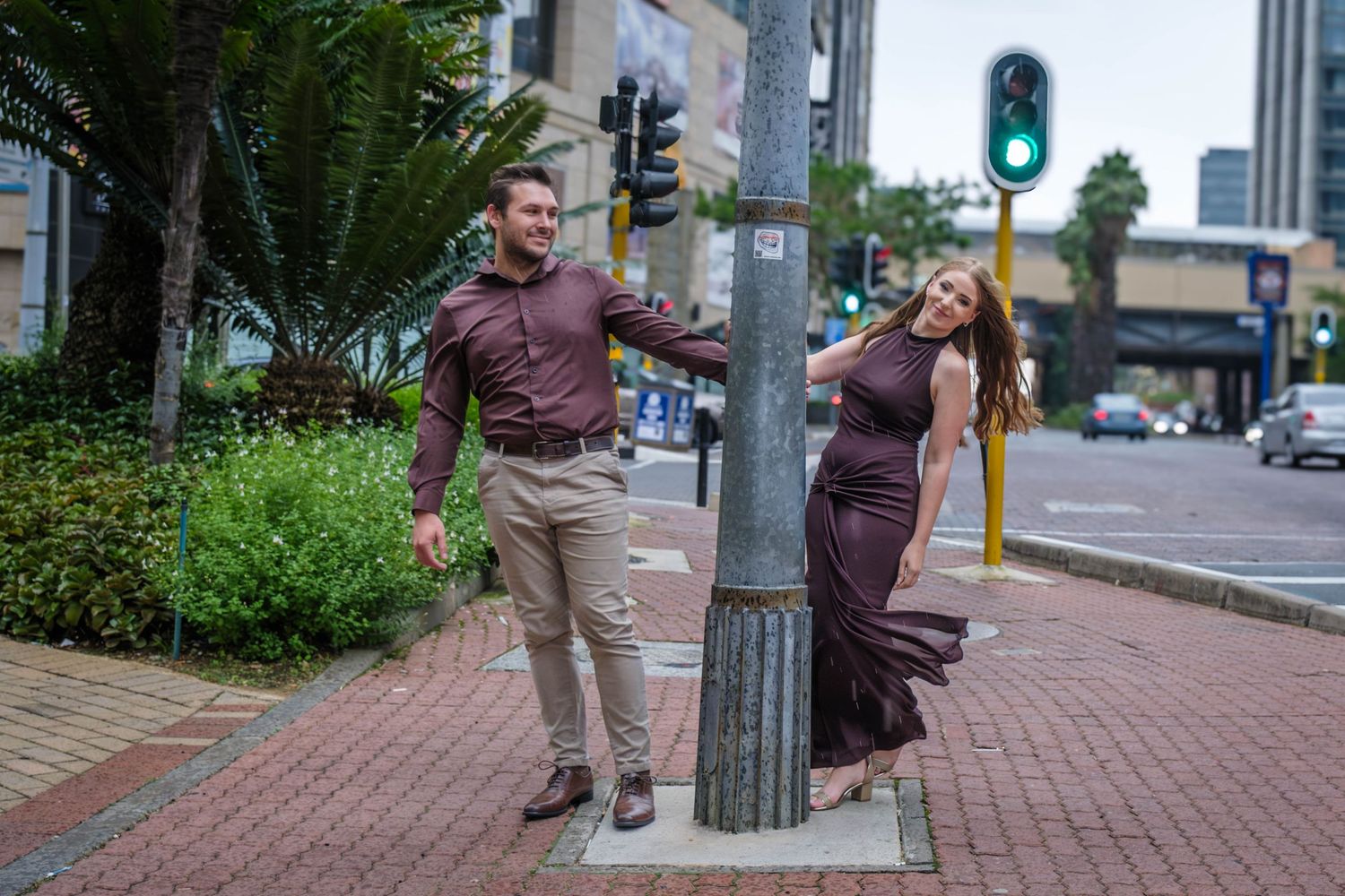 Stylish couple poses playfully around urban street pole in city center with green traffic light backdrop.