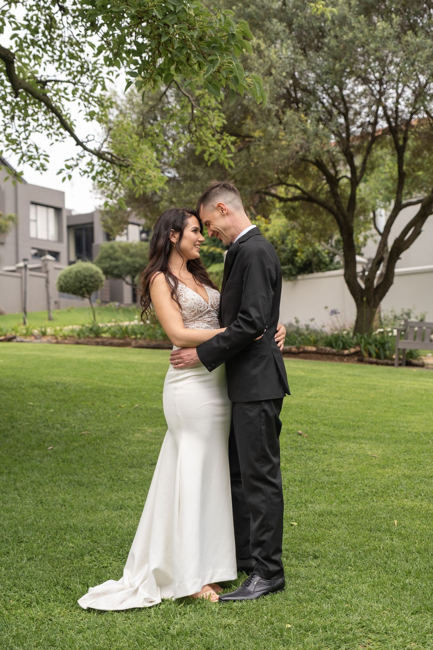 Bride and groom embracing forehead to forehead on green lawn surrounded by lush garden trees.
