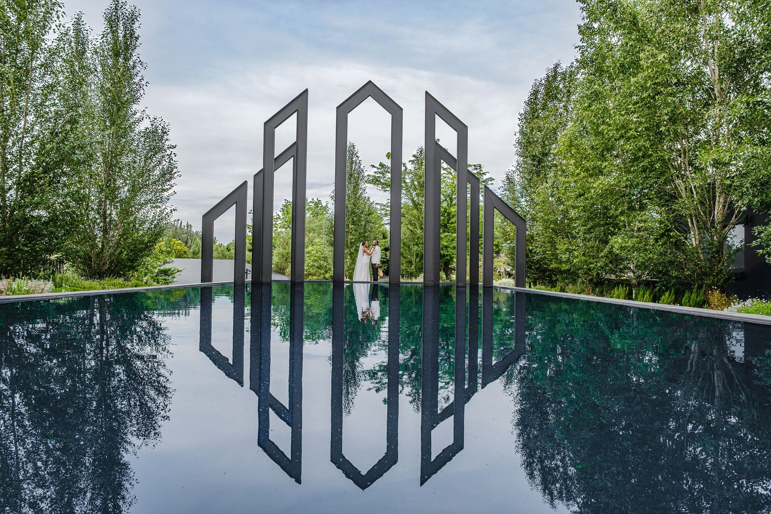 Bride and groom reflected in still pool beside geometric metal sculptures.