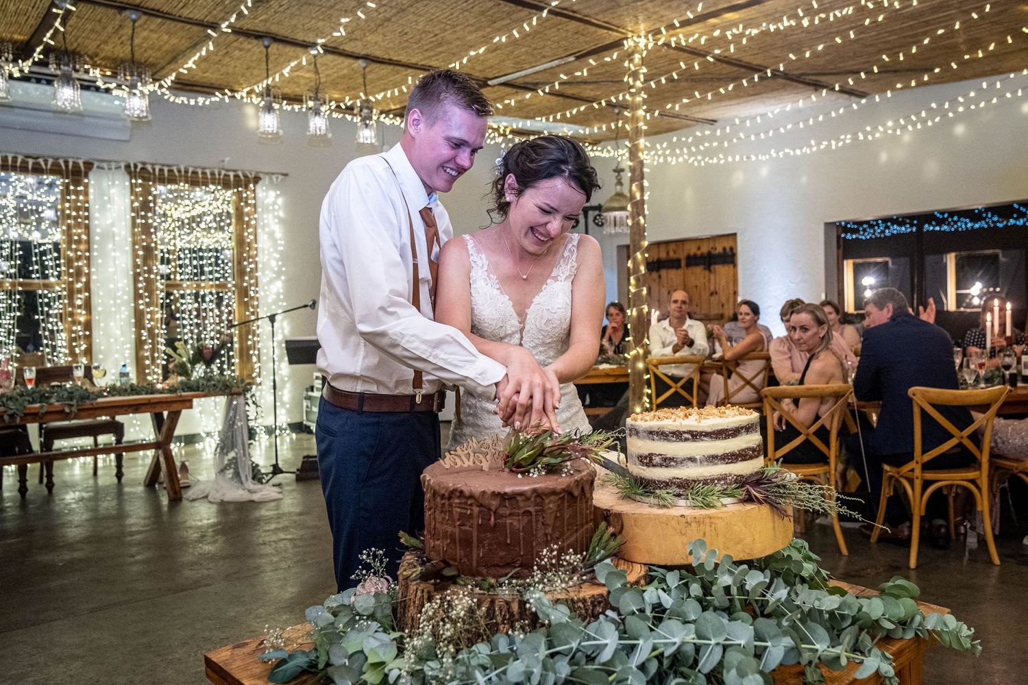 Bride and groom smile while cutting a rustic naked wedding cake at a fairy-light decorated reception.