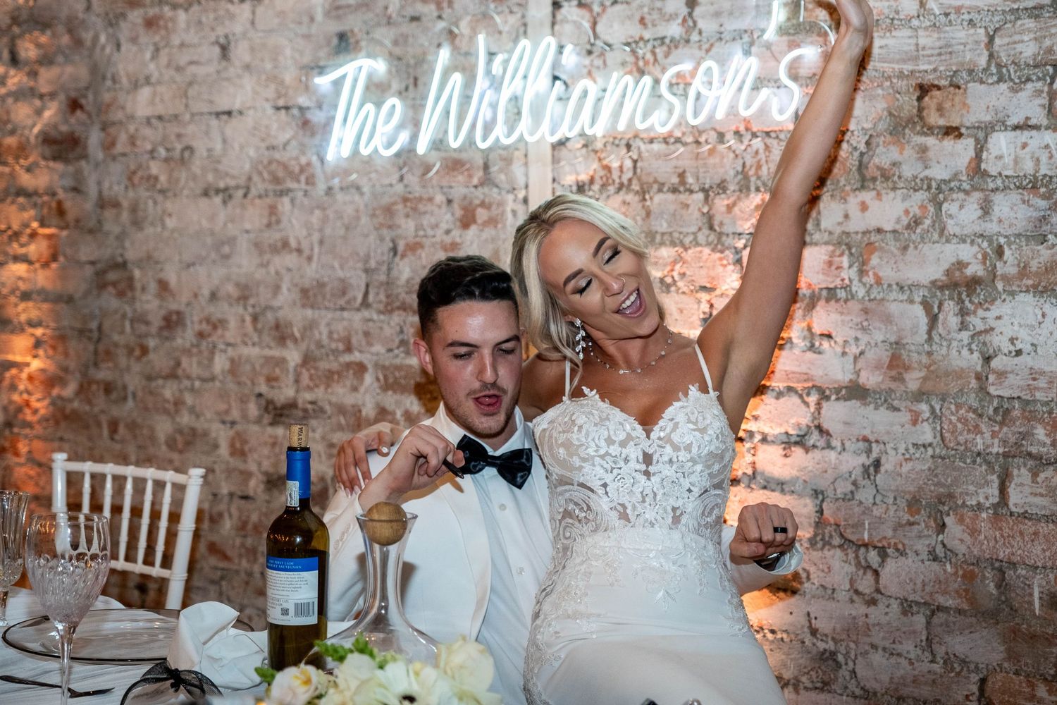 Joyful bride and groom celebrate at sweetheart table beneath The Williamsons neon sign.