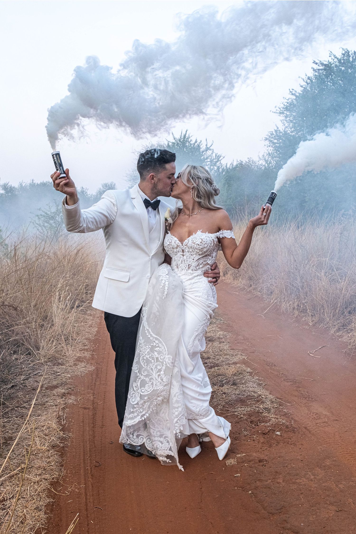 Bride and groom kissing while holding colorful smoke bombs on a rustic dirt road during wedding shoot.
