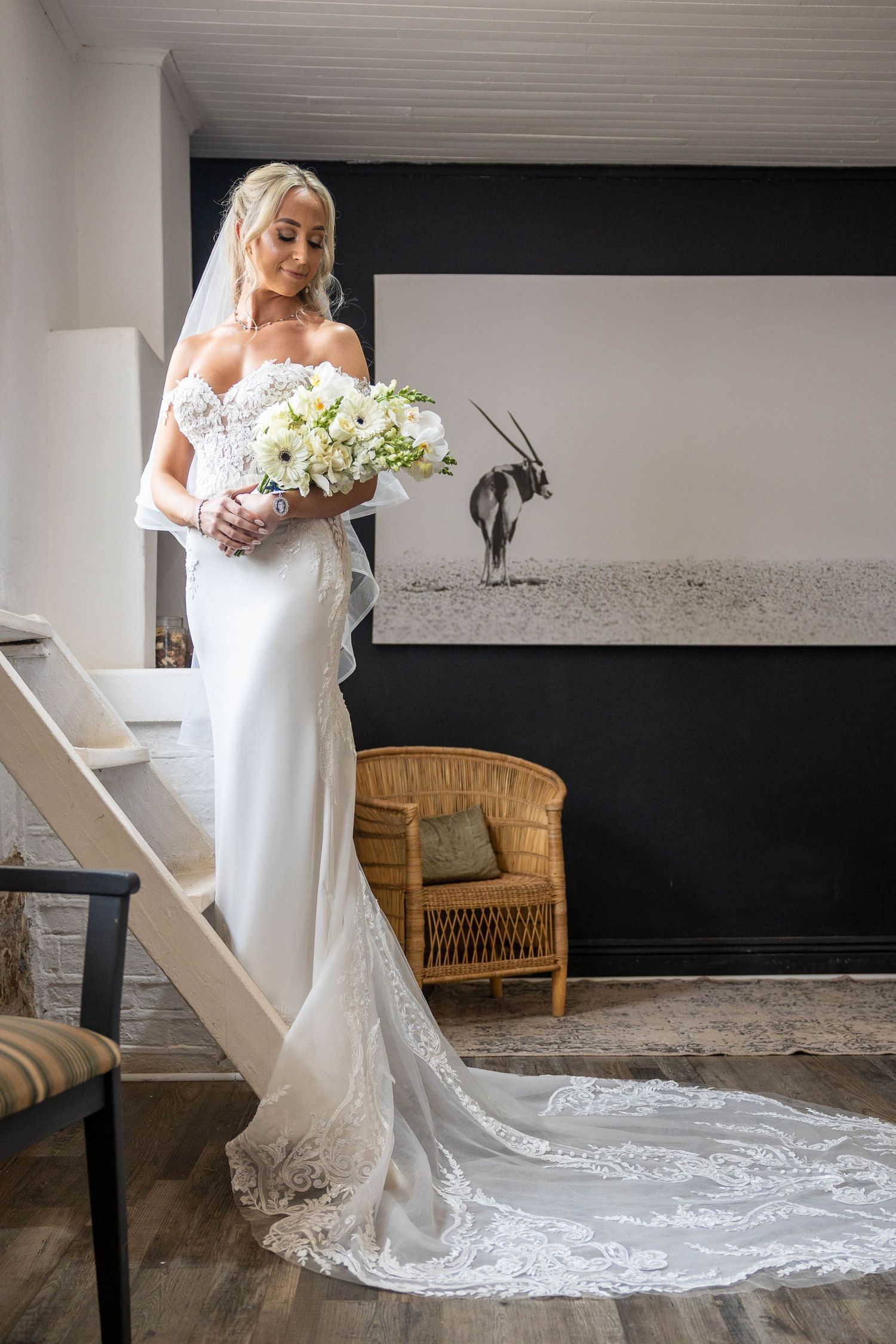 Elegant bride in off-shoulder lace gown holds white bouquet beside staircase in rustic interior.