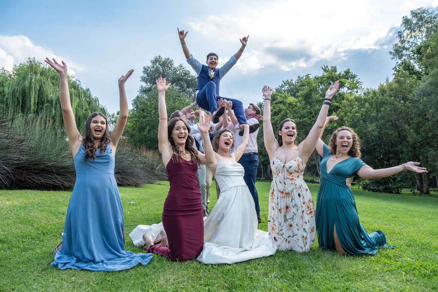 Excited wedding party celebrating with raised arms on green lawn.