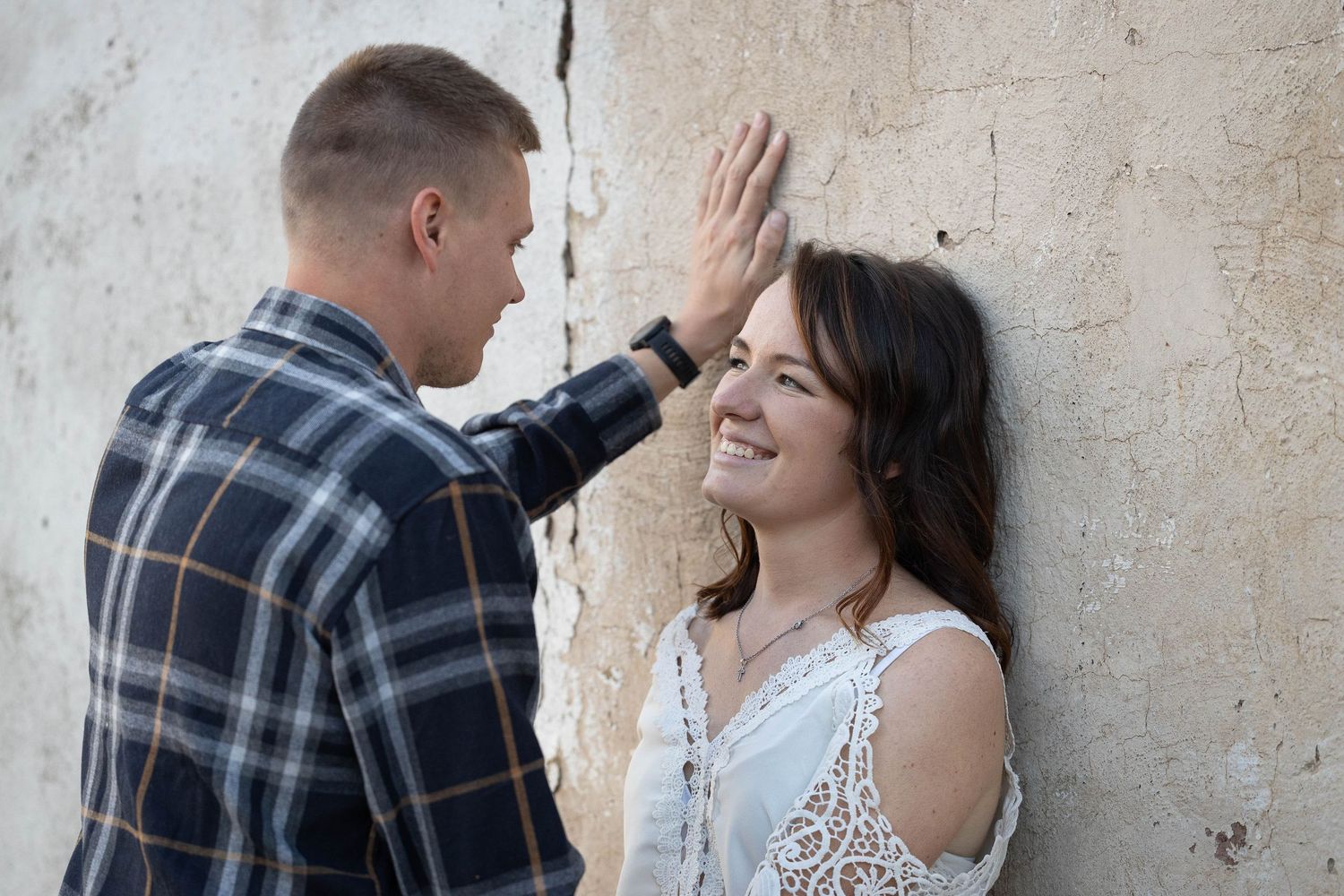 Man in plaid shirt placing hand on wall above laughing woman during romantic outdoor engagement photo session.