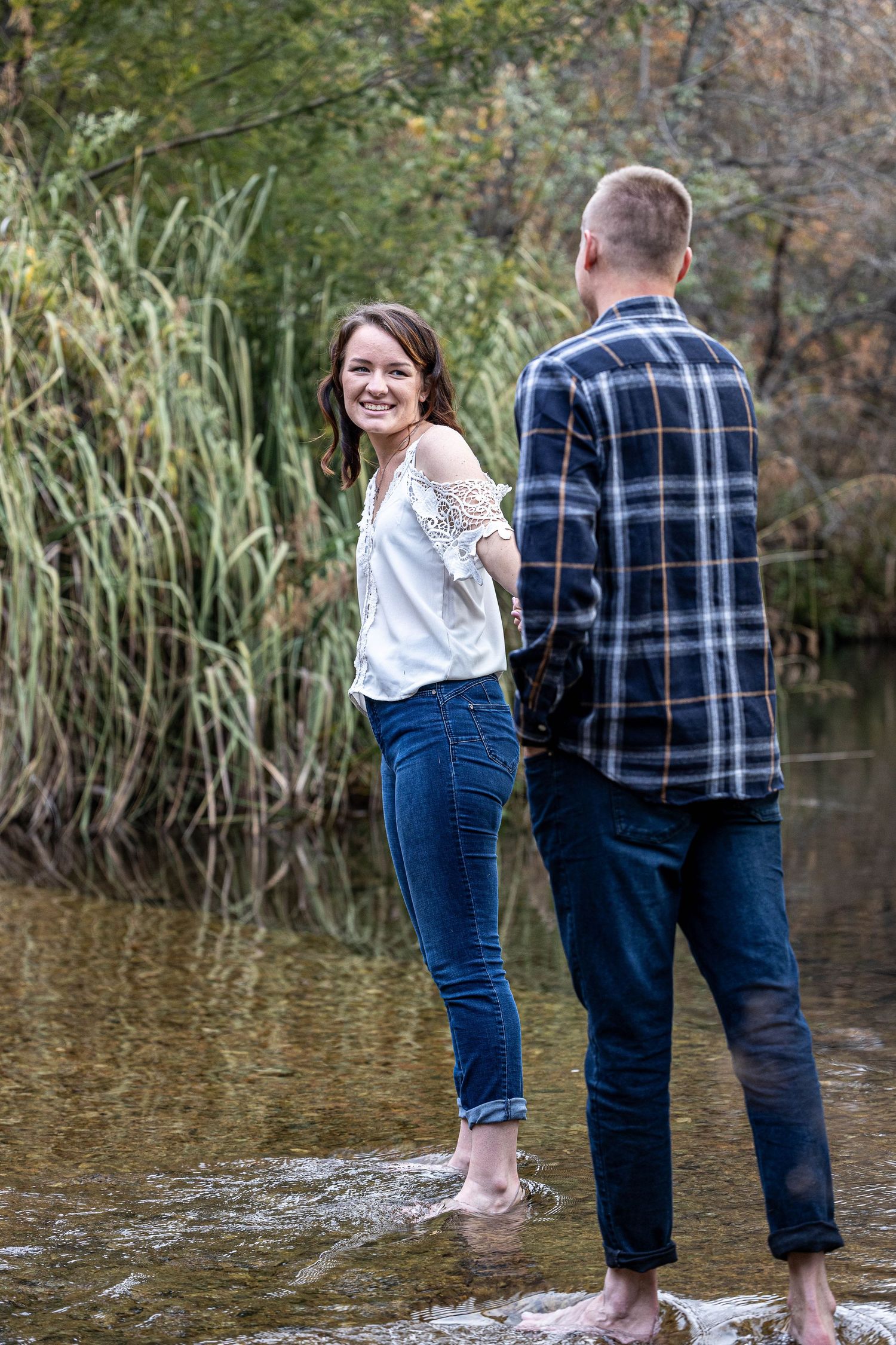 Smiling woman in white top and jeans holds hands with man while wading barefoot in a shallow creek.