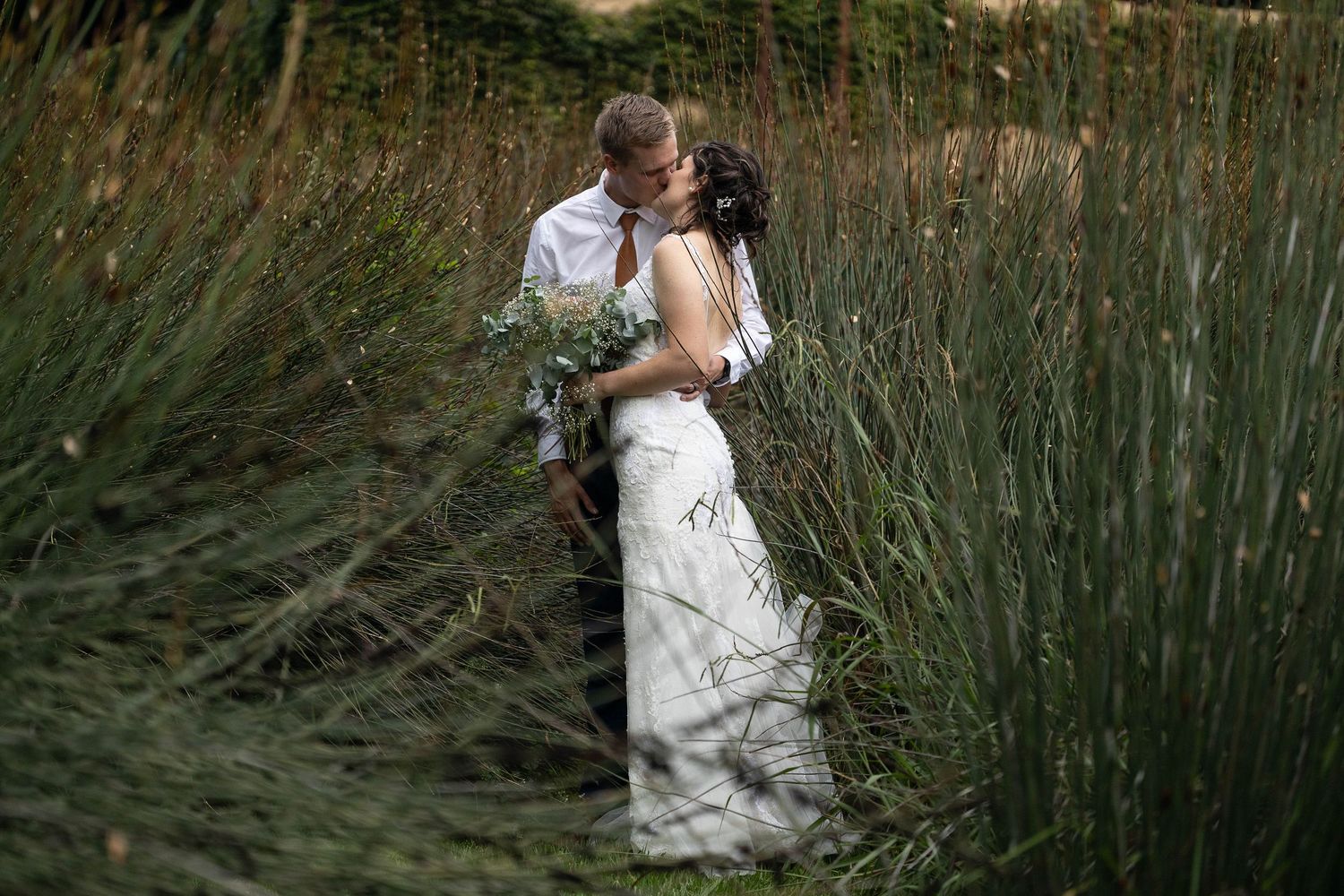 Wedding couple embracing and kissing among tall green grass with floral bouquet.