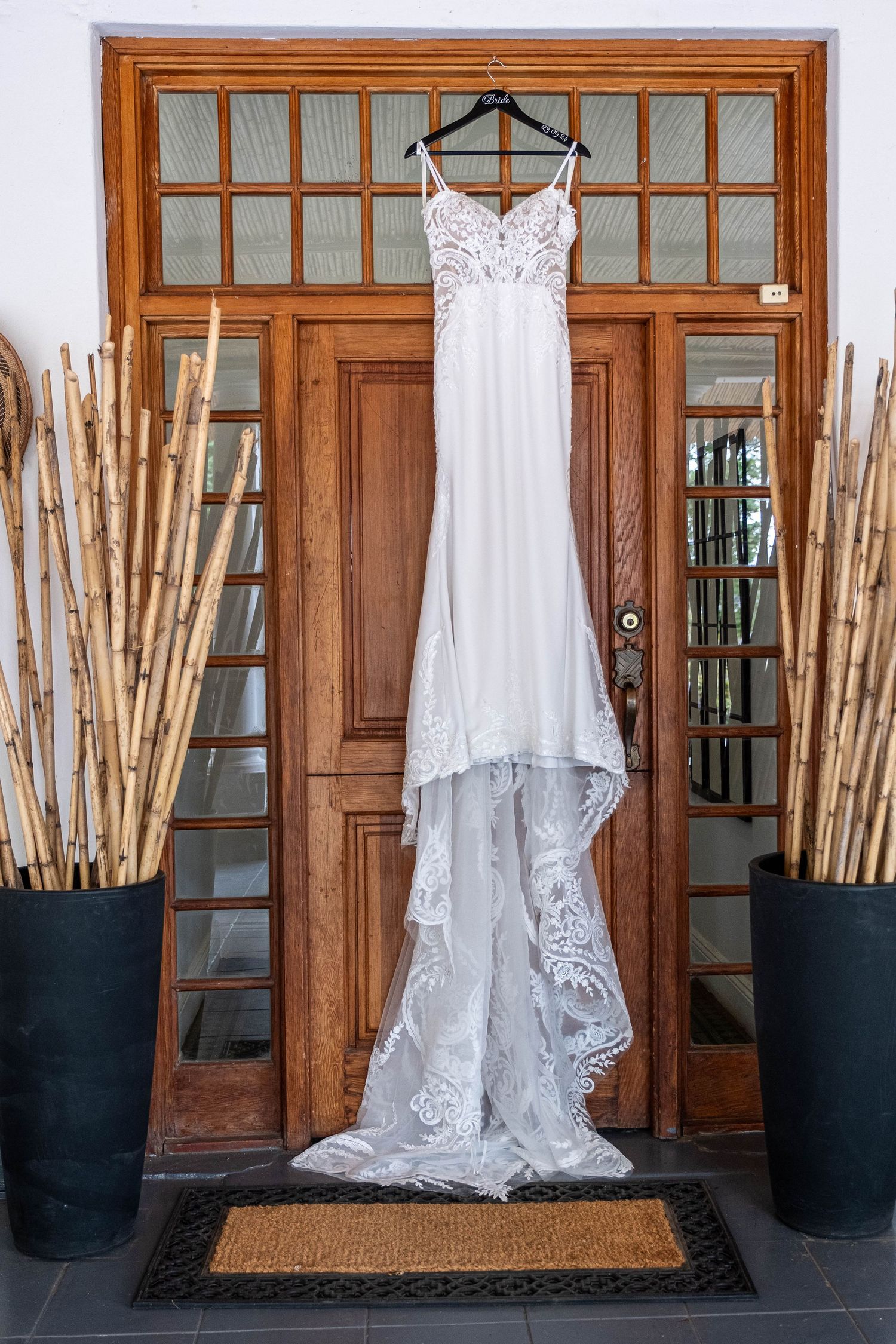 Elegant white lace wedding dress hanging on wooden door with bamboo plants on either side.