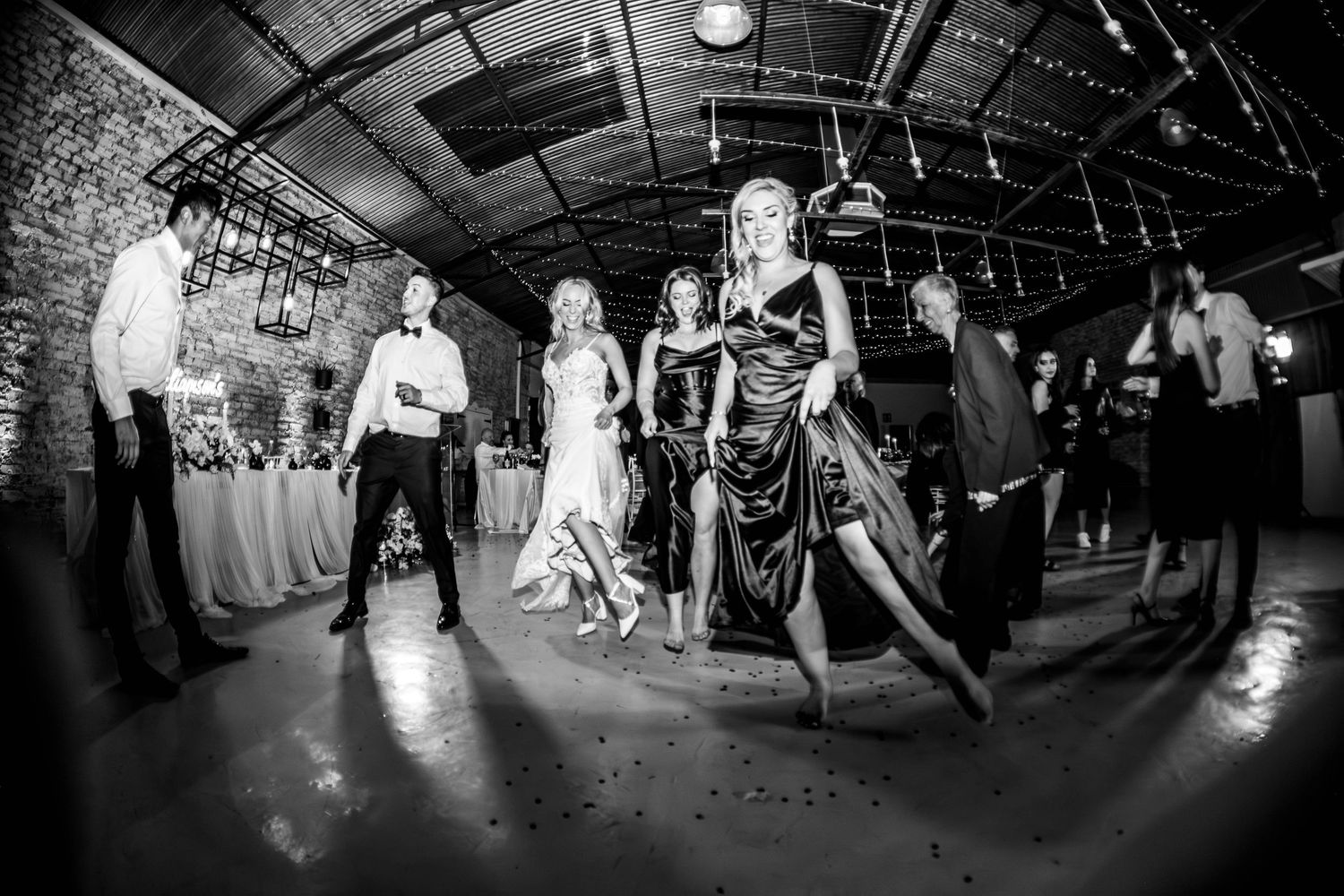 Black and white photo of elegantly dressed wedding guests walking across a rustic reception dance floor.