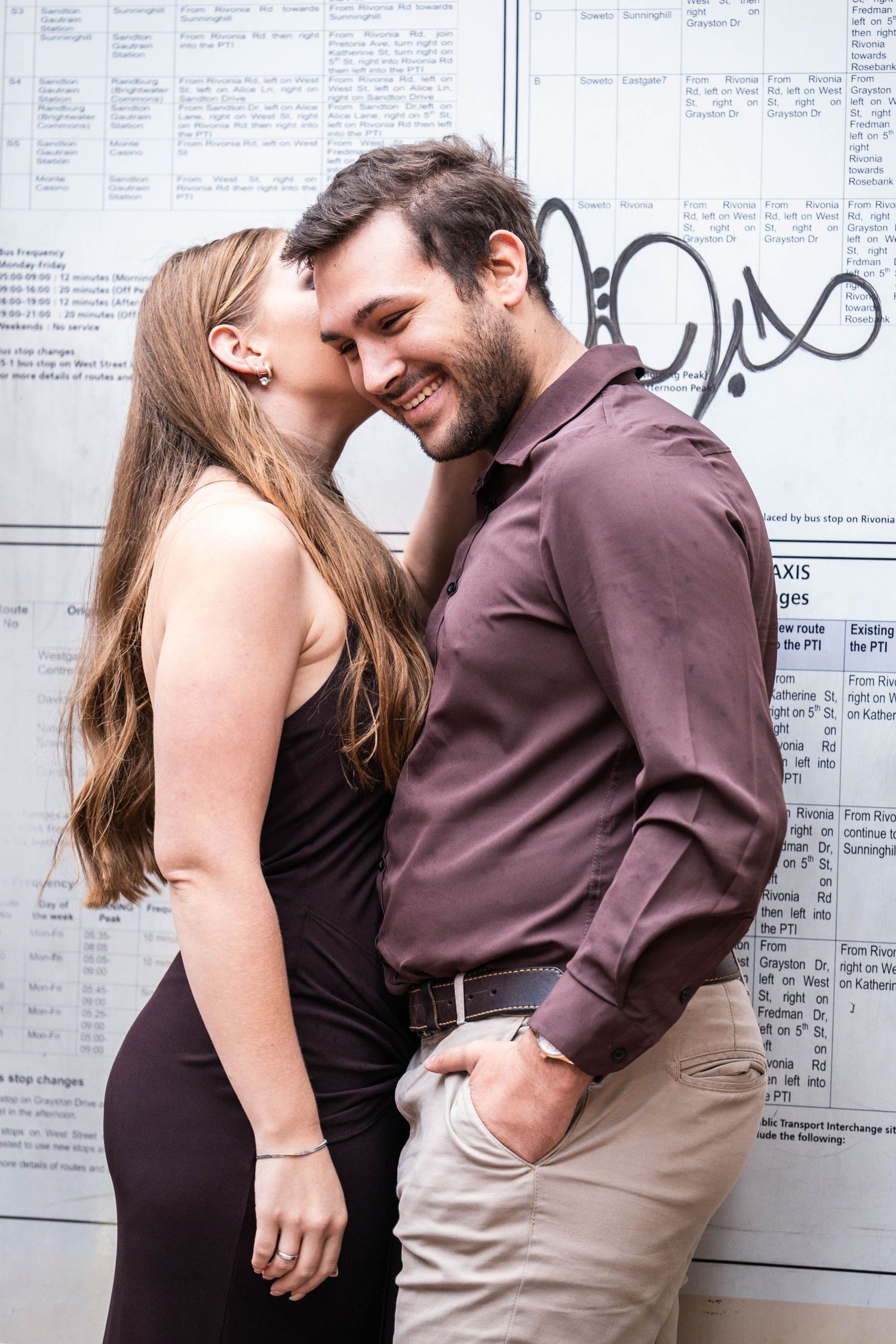 Smiling couple shares tender kiss moment against newspaper covered wall, woman in black dress kissing man's cheek.