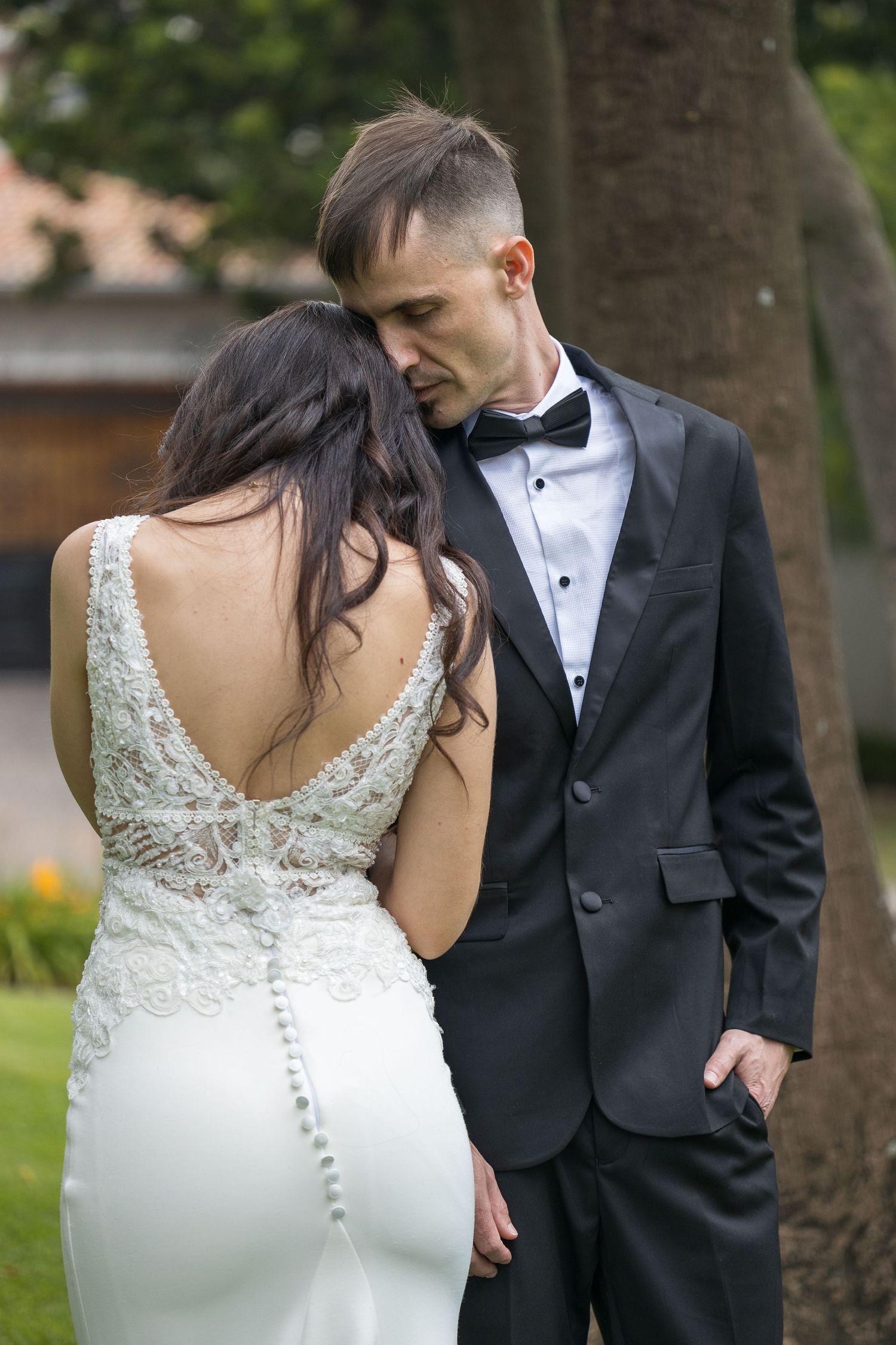 Groom in black tuxedo tenderly kissing brides head while she shows lace back wedding gown.