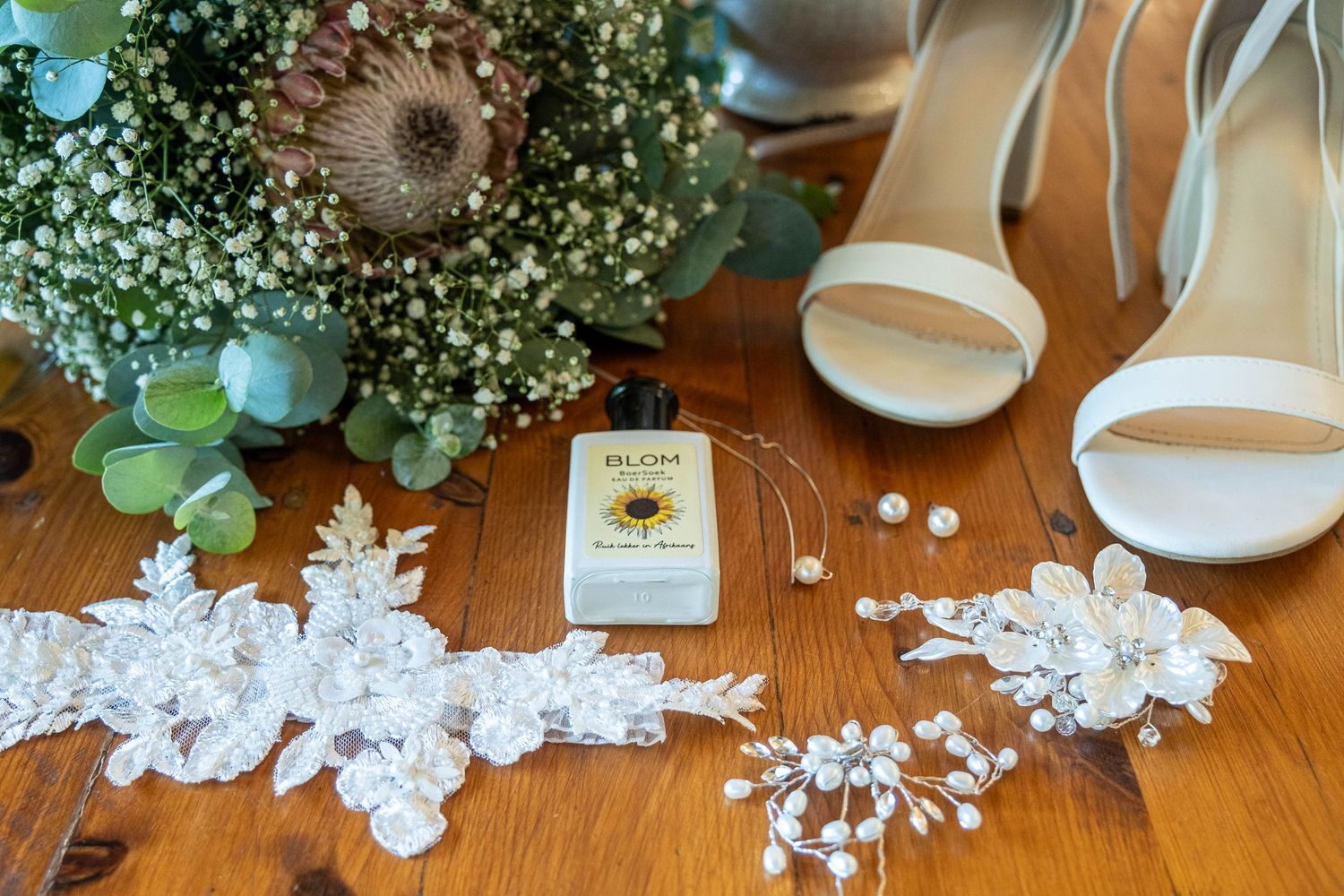 Bridal flat lay featuring white heels, lace garter, pearl jewelry, perfume, and a protea bouquet on wood.