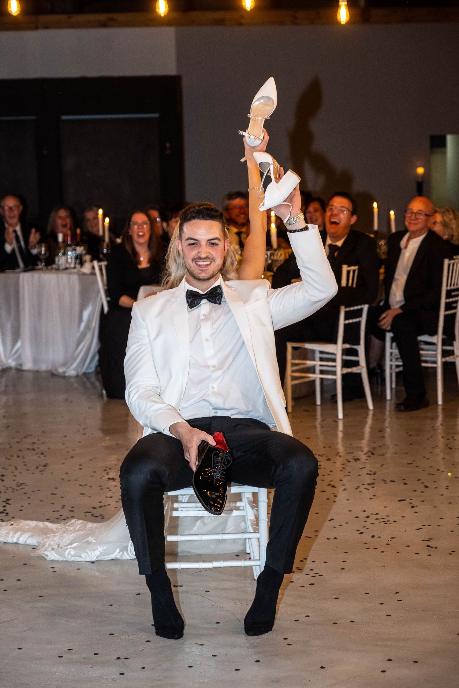Smiling groom in white tuxedo holds up brides shoe during wedding shoe game at reception.