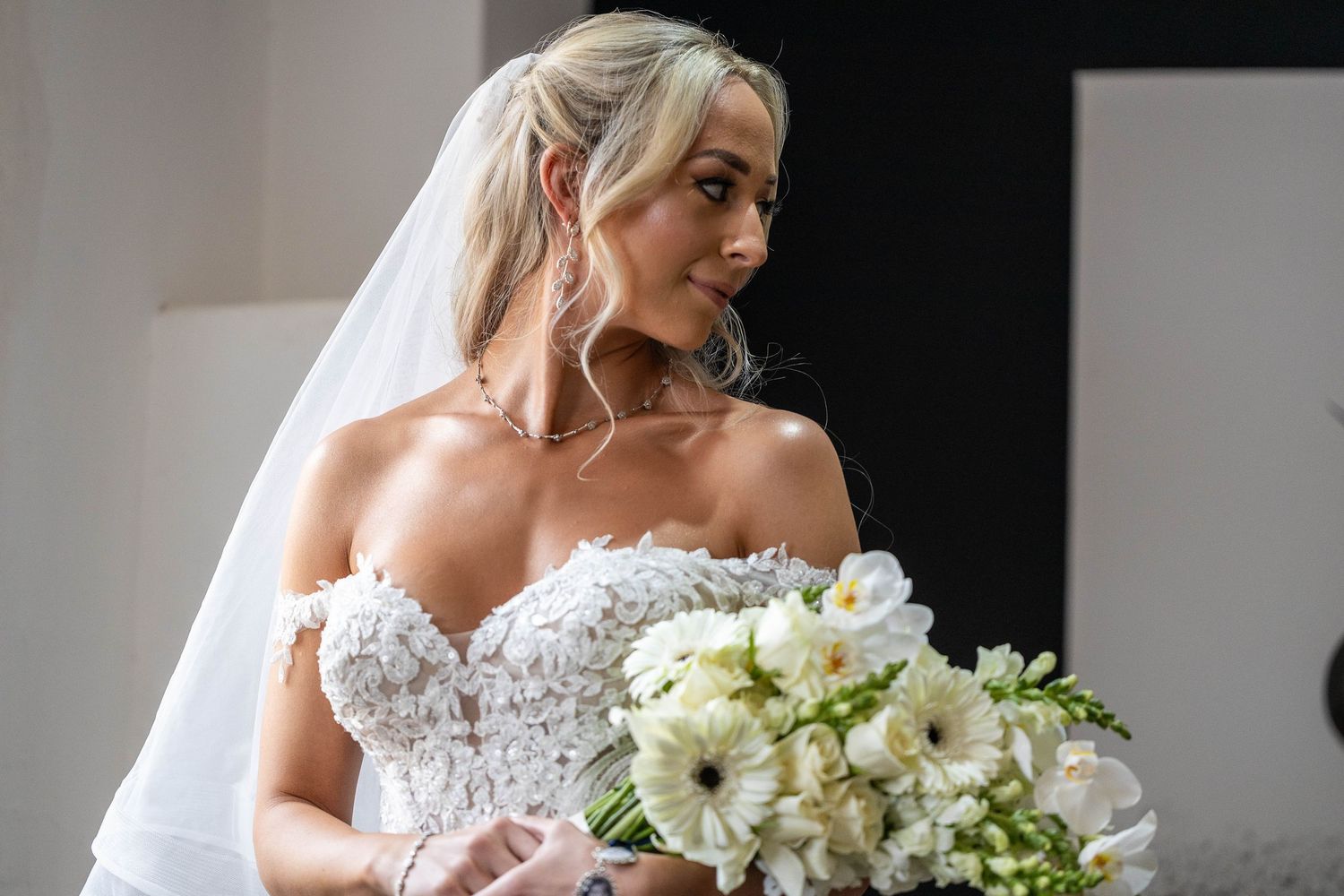Beautiful bride in off-shoulder lace gown with veil holds a lush white floral bouquet.