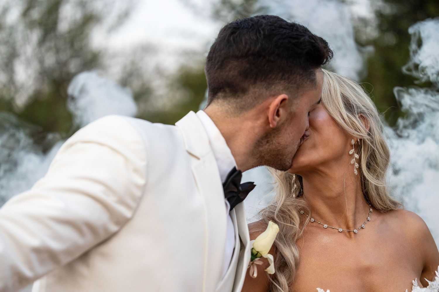 Close-up of bride and groom sharing a passionate kiss surrounded by romantic white smoke outdoors.