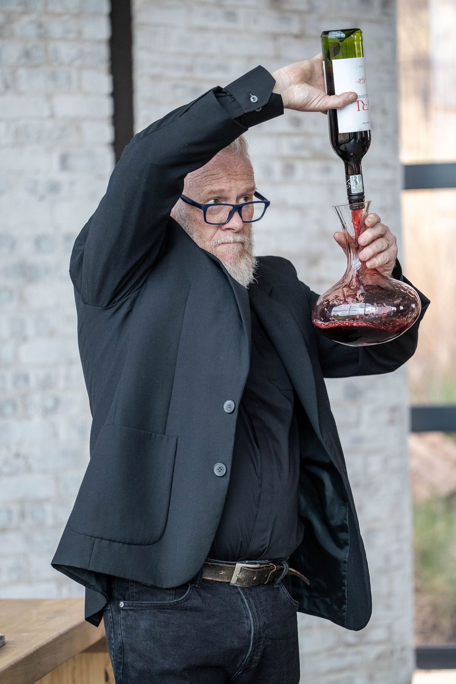 Elderly man in black suit dramatically decants red wine into glass decanter.