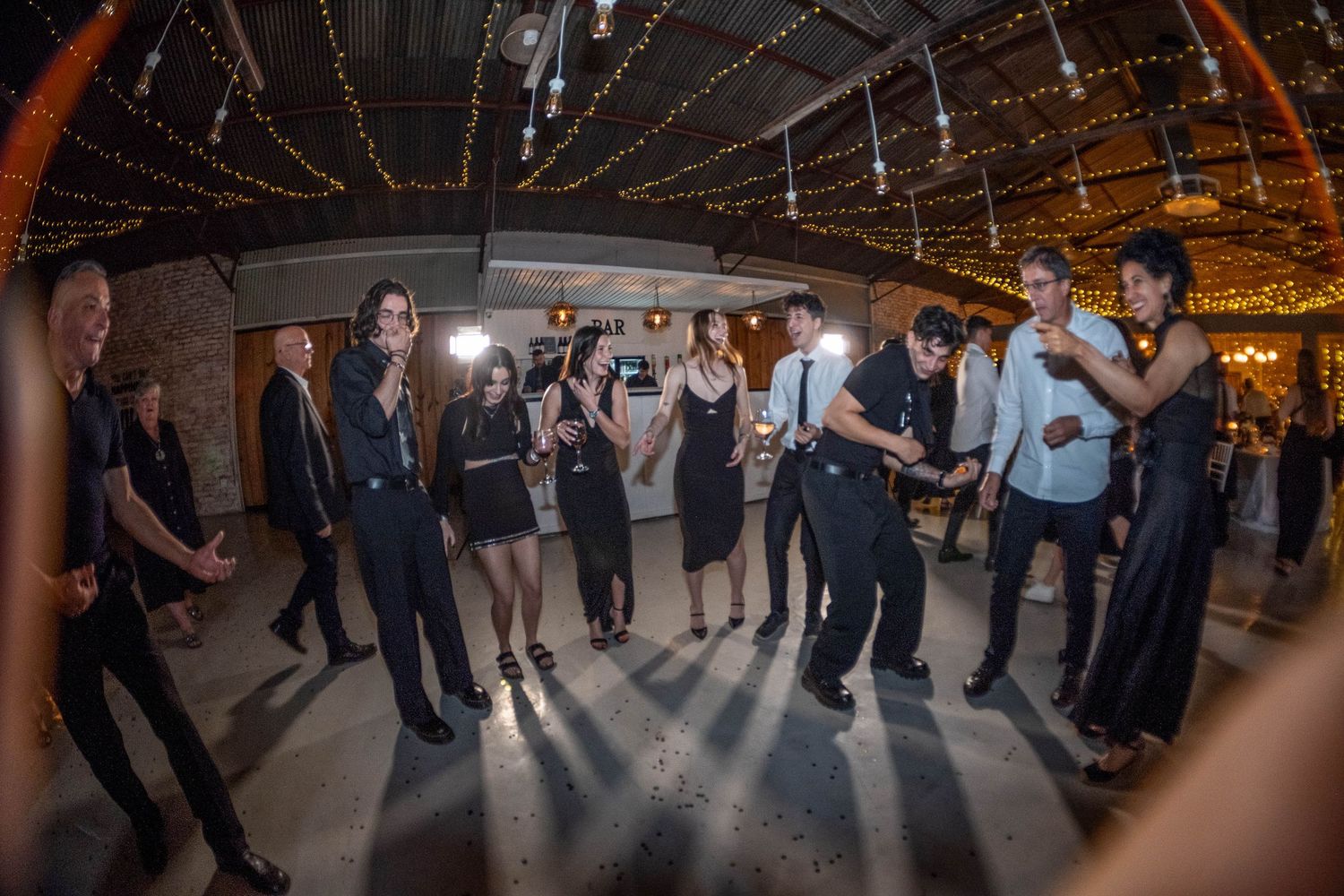 Wedding guests dancing in circle on dance floor at indoor reception with string lights overhead.