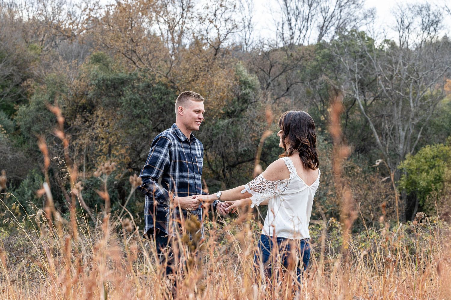 Couple holds hands while walking through tall golden grass field with autumn trees behind them.