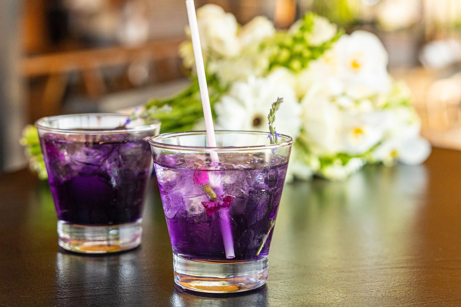 Two vibrant purple cocktails with ice being poured in glasses on a dark table with white flowers.