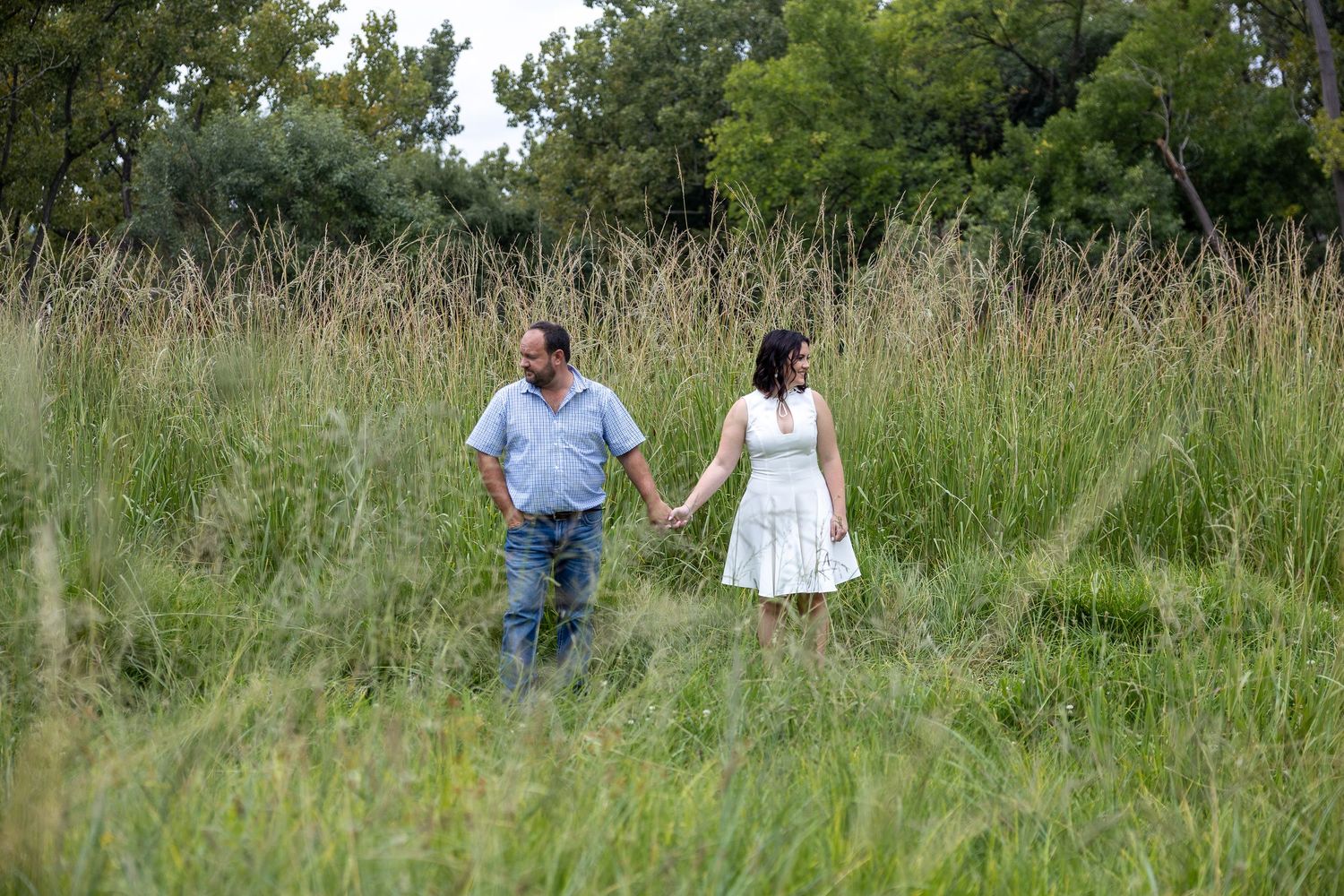 Couple holding hands walking through tall grass in a lush green meadow.