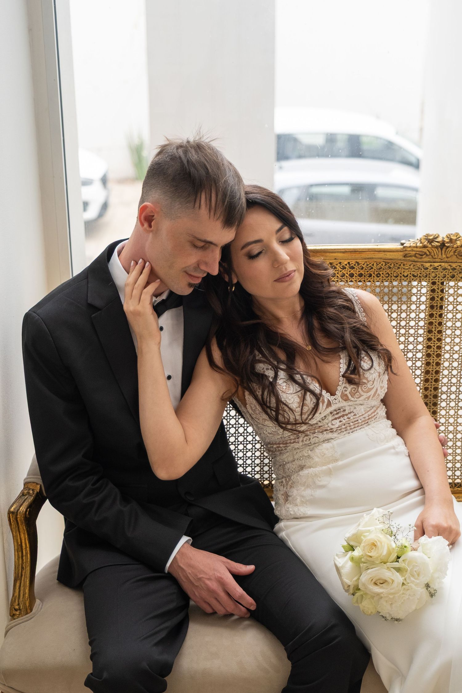 A bride and groom in wedding attire sit closely together on a gold ornate bench, sharing a tender moment.