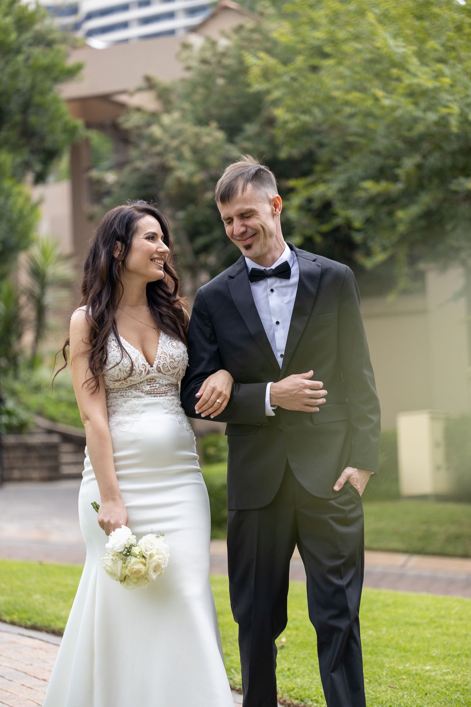 Happy bride and groom walking together in garden, bride holding white bouquet in elegant wedding attire.