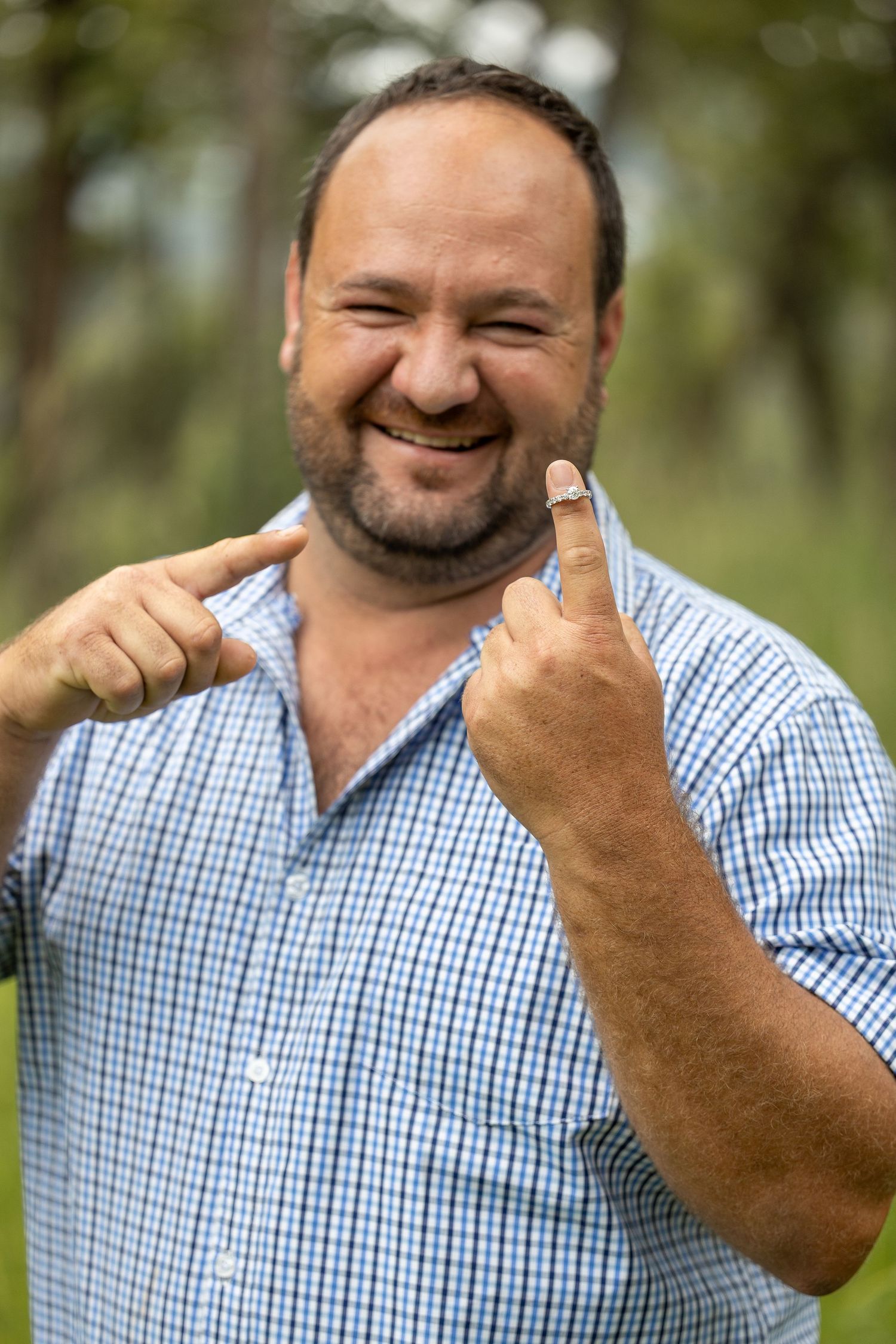 Smiling bearded man in blue plaid shirt pointing to his ring finger outdoors.