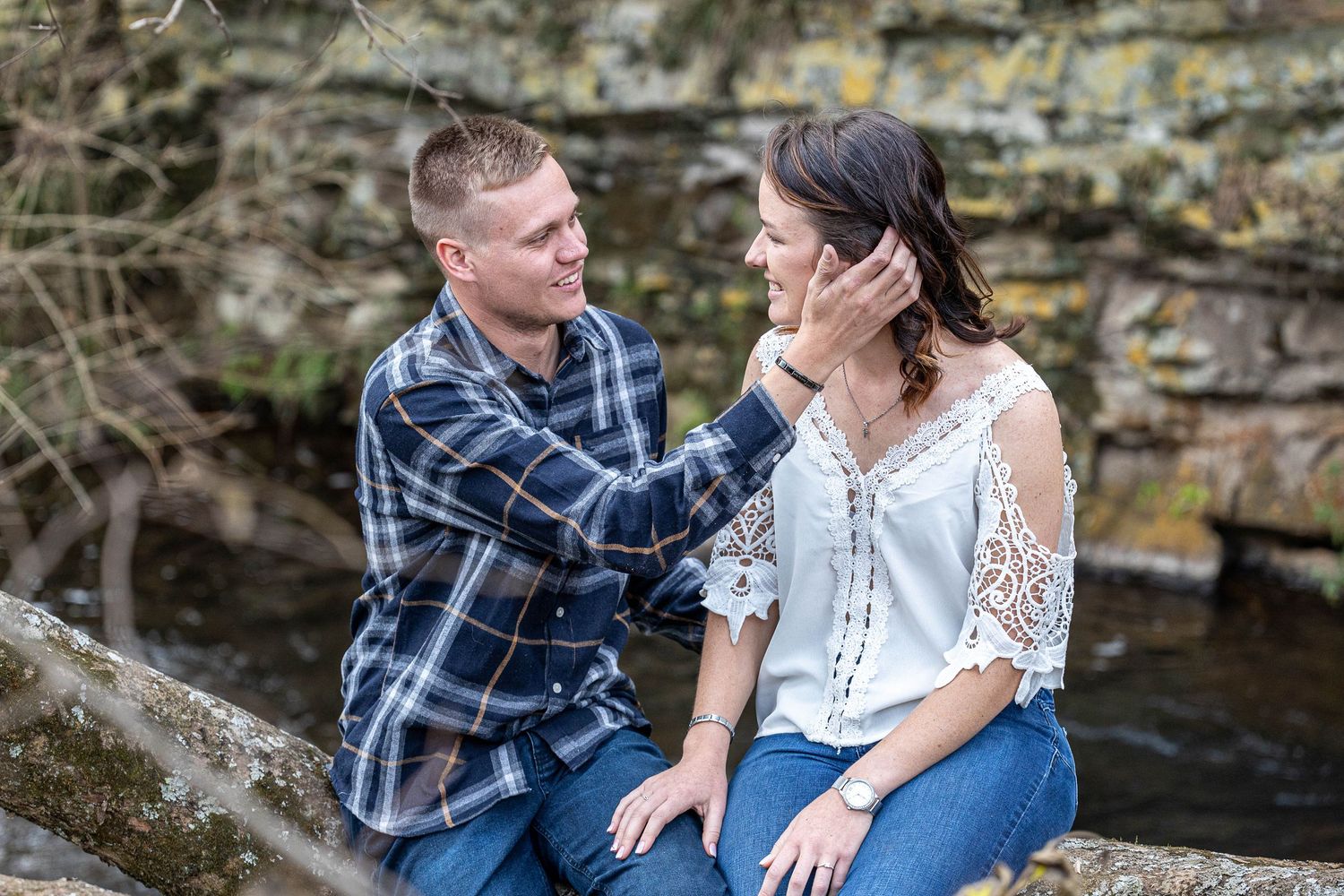 Couple sitting on rocks by a creek, man tenderly touching woman's face during engagement photo session.