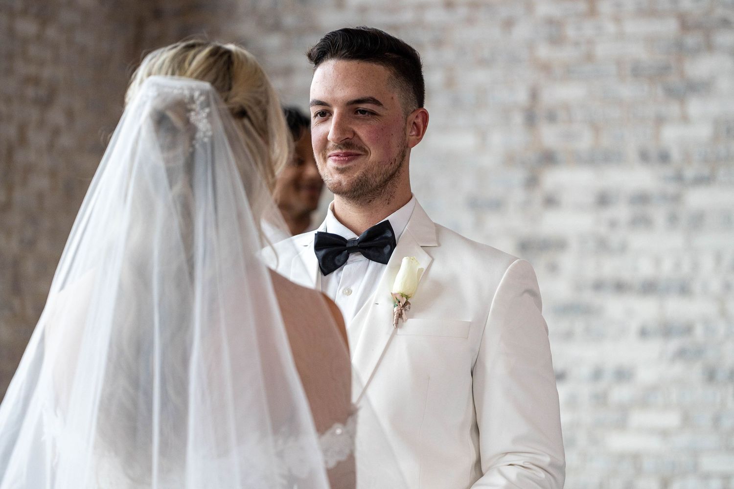 A handsome groom in a white jacket and black bow tie smiles warmly at his bride during the ceremony.