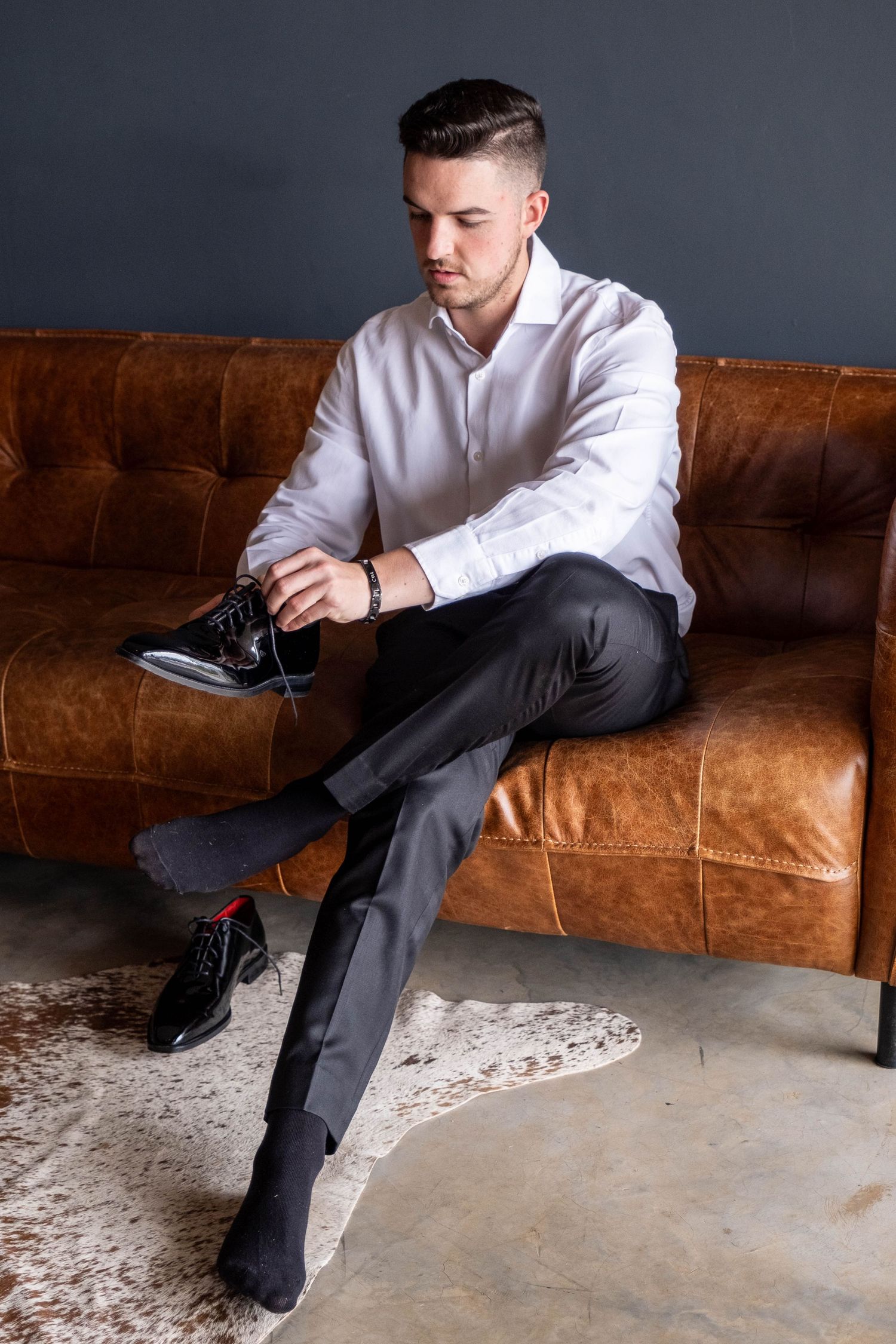 Young man in white shirt lacing up black Oxford shoes on leather sofa.