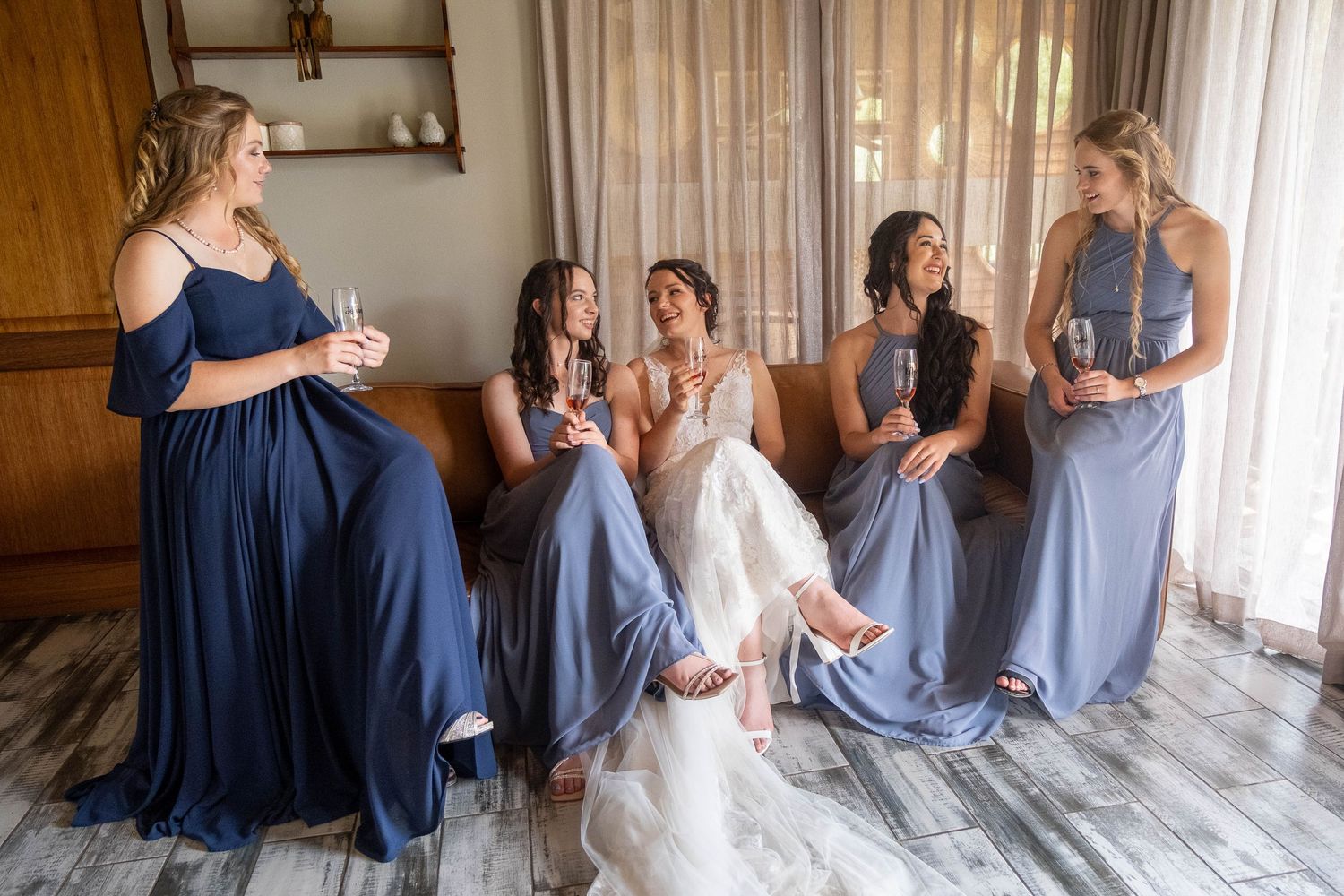 Bride and four bridesmaids in blue gowns sitting together sharing champagne before the wedding ceremony.