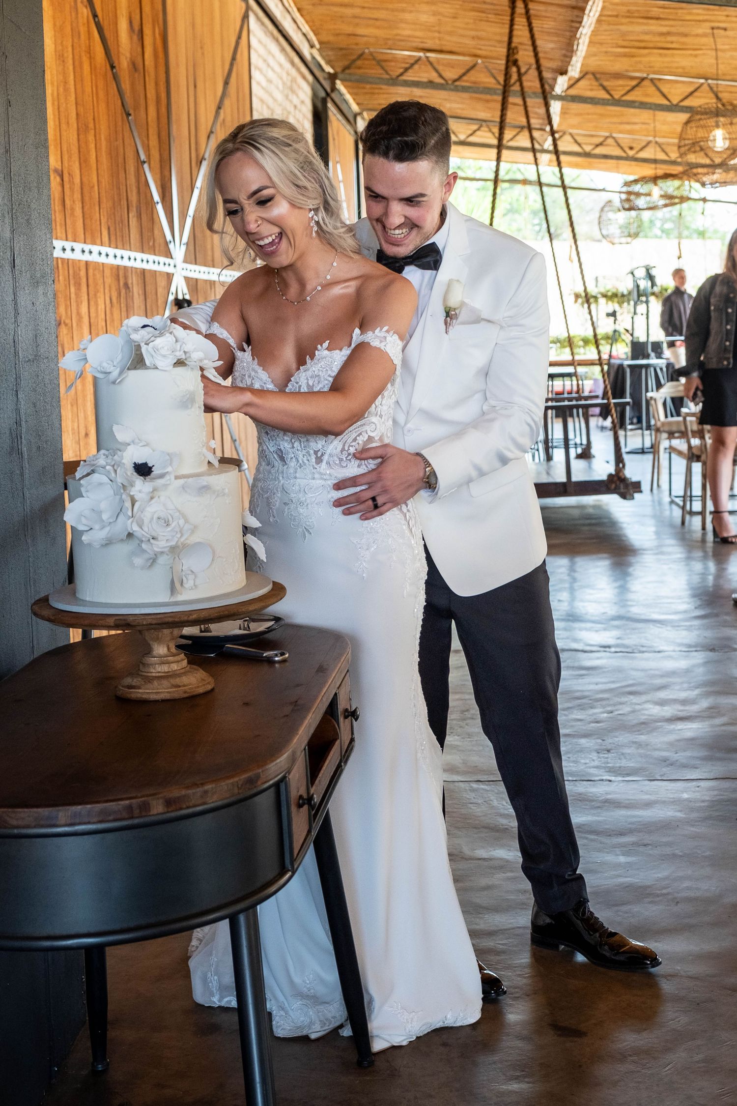 Bride and groom cutting elegant white floral wedding cake together at rustic barn reception venue.