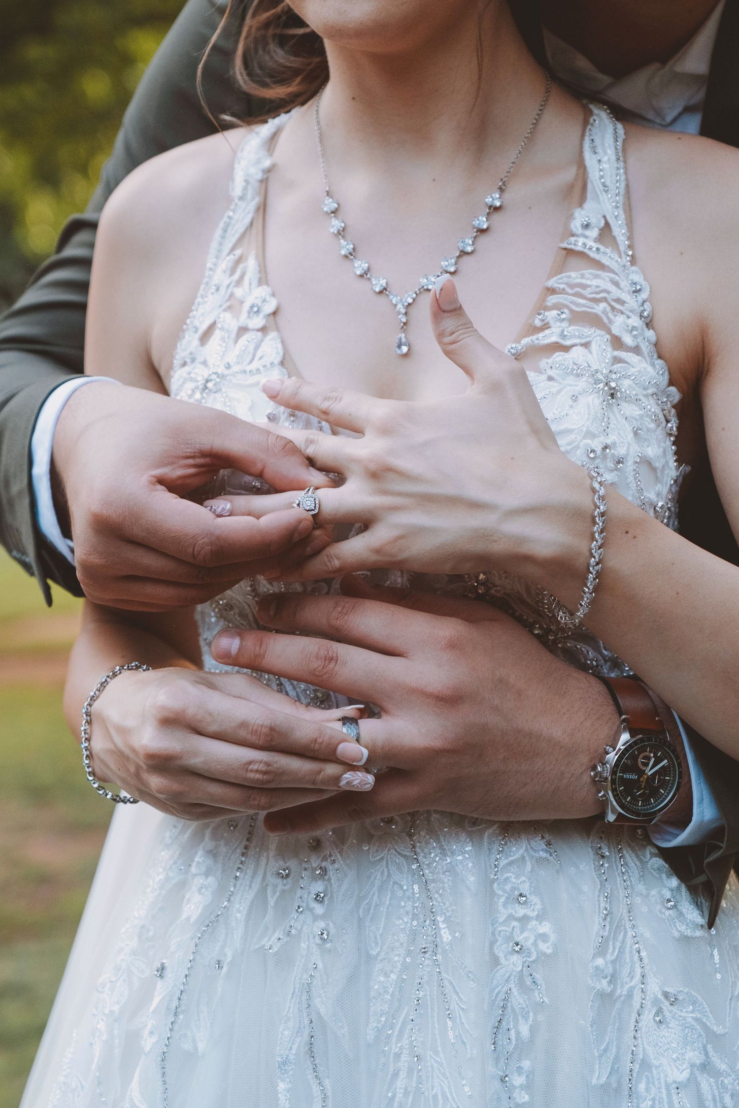 Bride and groom holding hands showing wedding rings, bride wearing lace dress and diamond necklace.