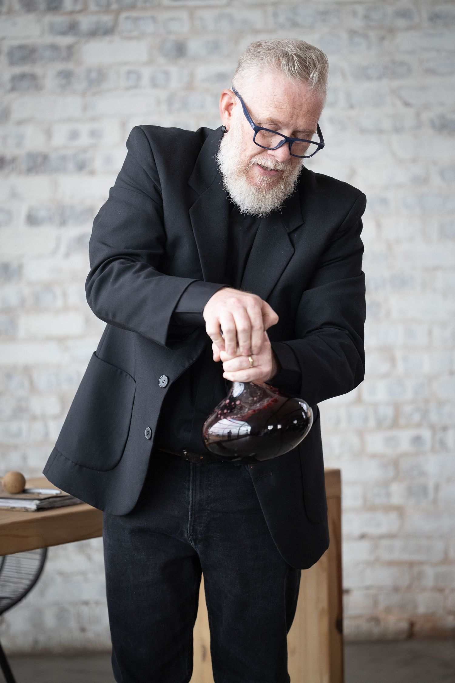 Bearded older man in black suit carefully handles wine decanter at wedding reception.