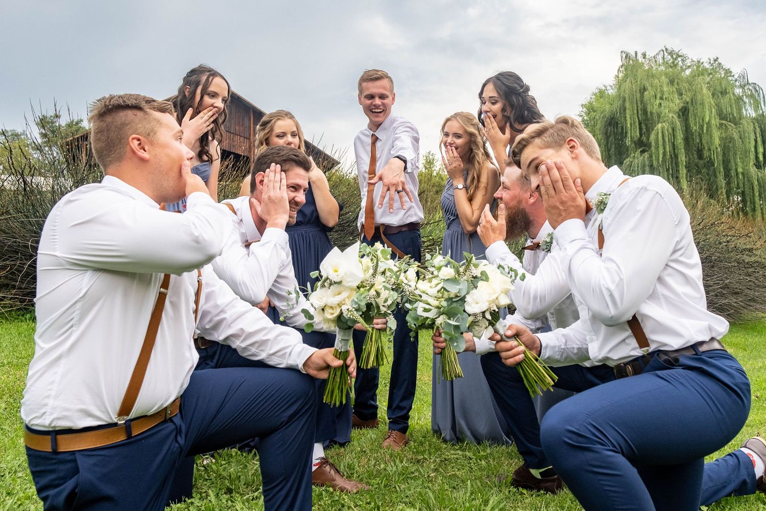 Wedding party poses playfully with groomsmen kneeling and everyone covering their faces in a garden.
