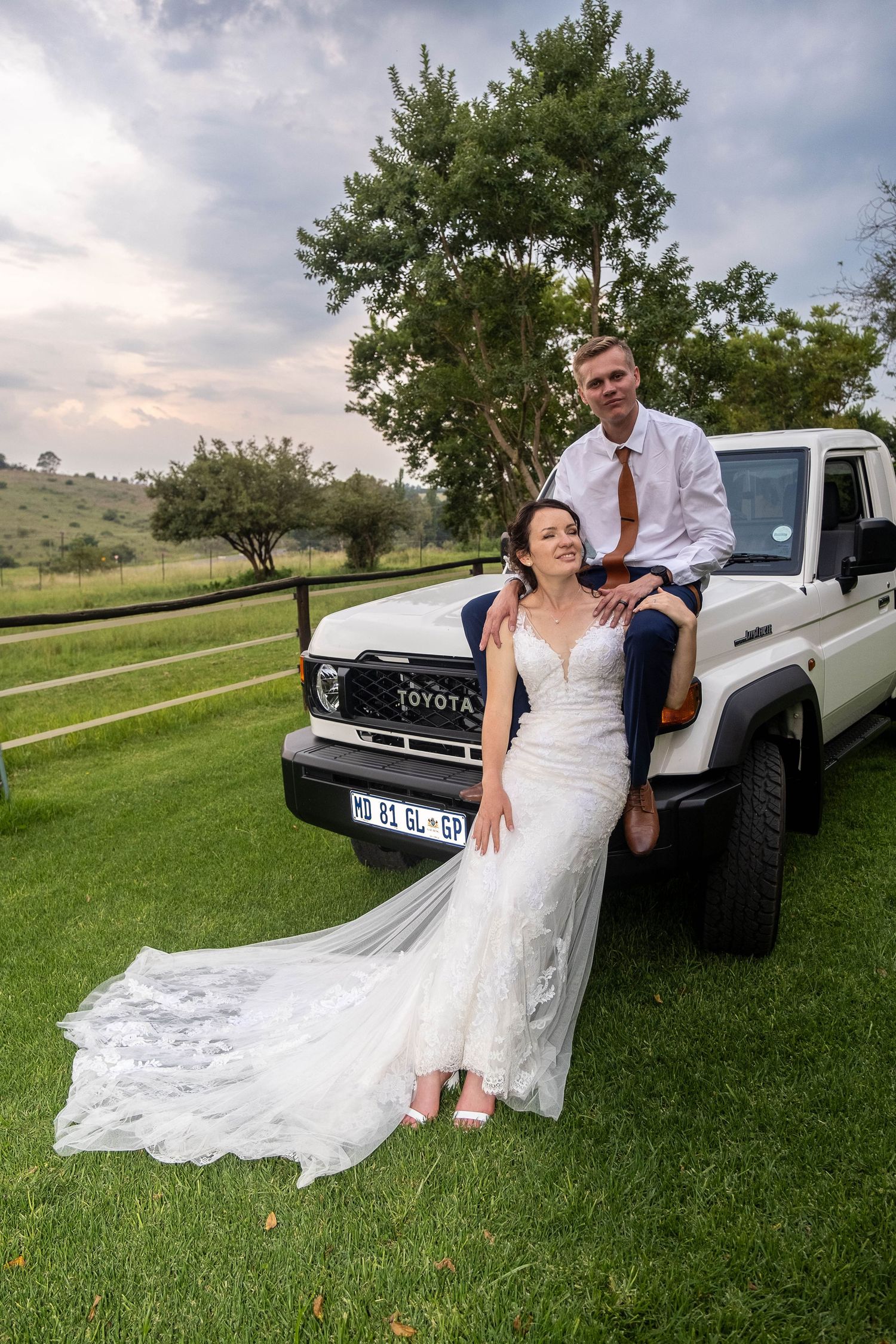 Bride in lace gown and groom in navy suit pose together by a white Toyota Land Cruiser on green grass.