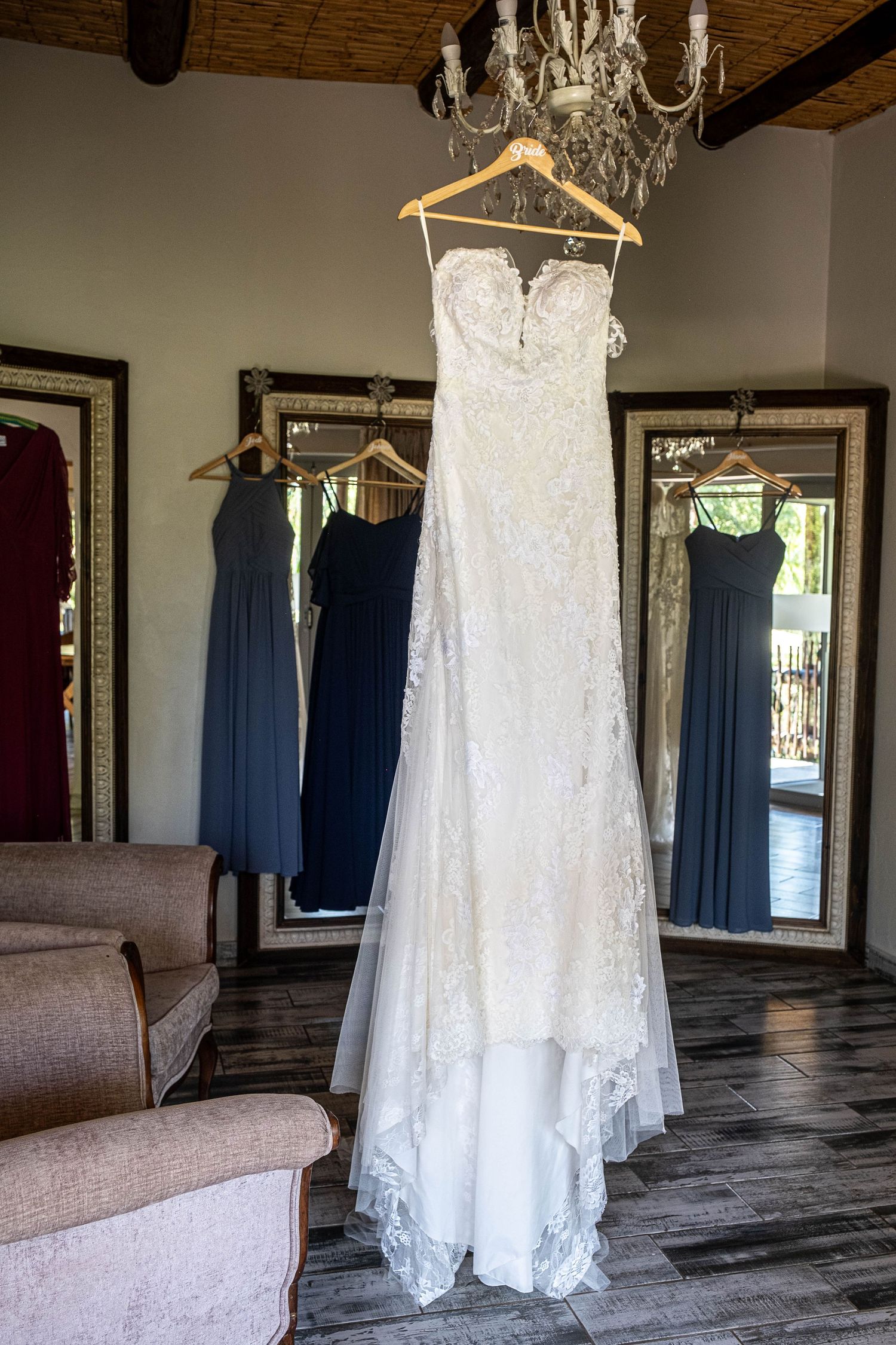 Elegant lace wedding dress hanging from a chandelier with navy blue bridesmaids dresses reflected in mirrors.