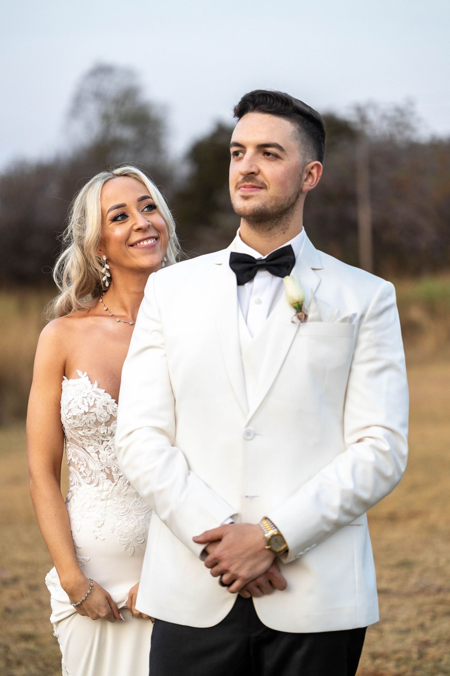 Smiling bride and groom posing outdoors, groom in white tuxedo with black bow tie and boutonniere.