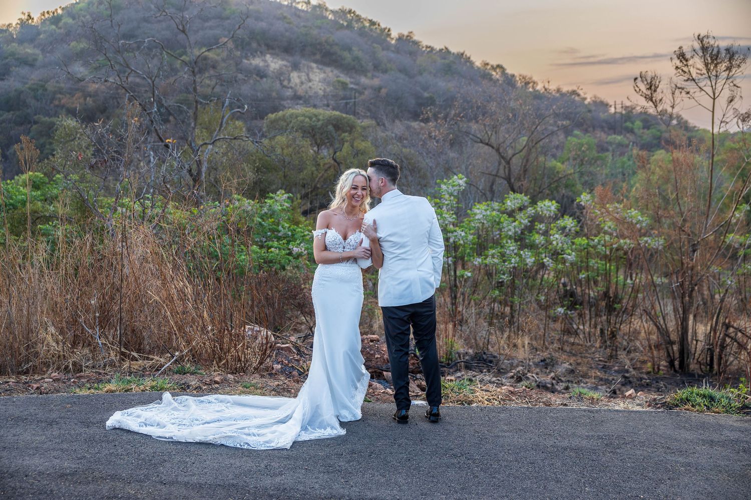 Wedding couple sharing a romantic moment on a road with a scenic mountain backdrop at sunset.