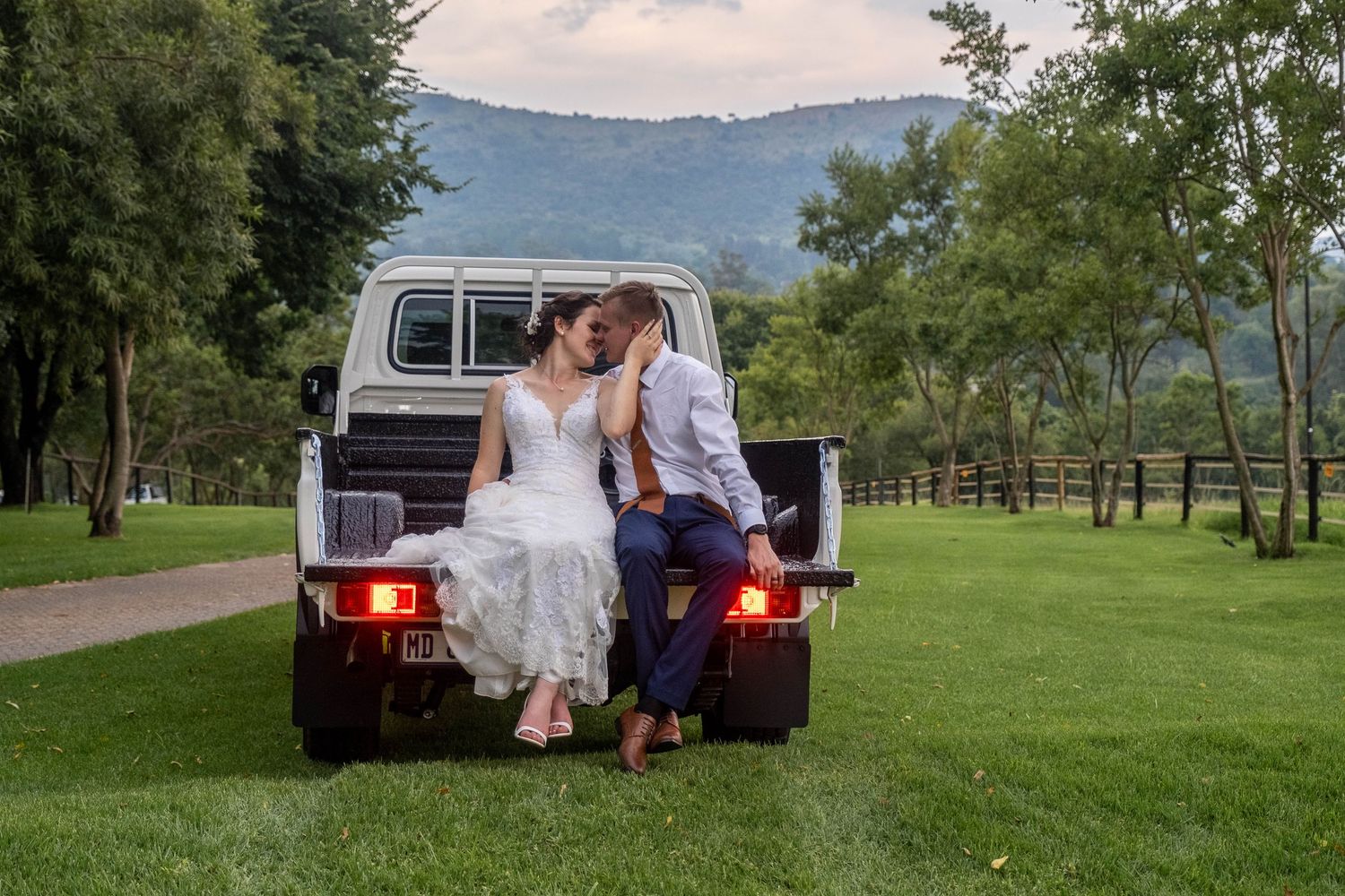 Bride and groom share a romantic kiss seated on the back of a white vintage truck in a scenic landscape.