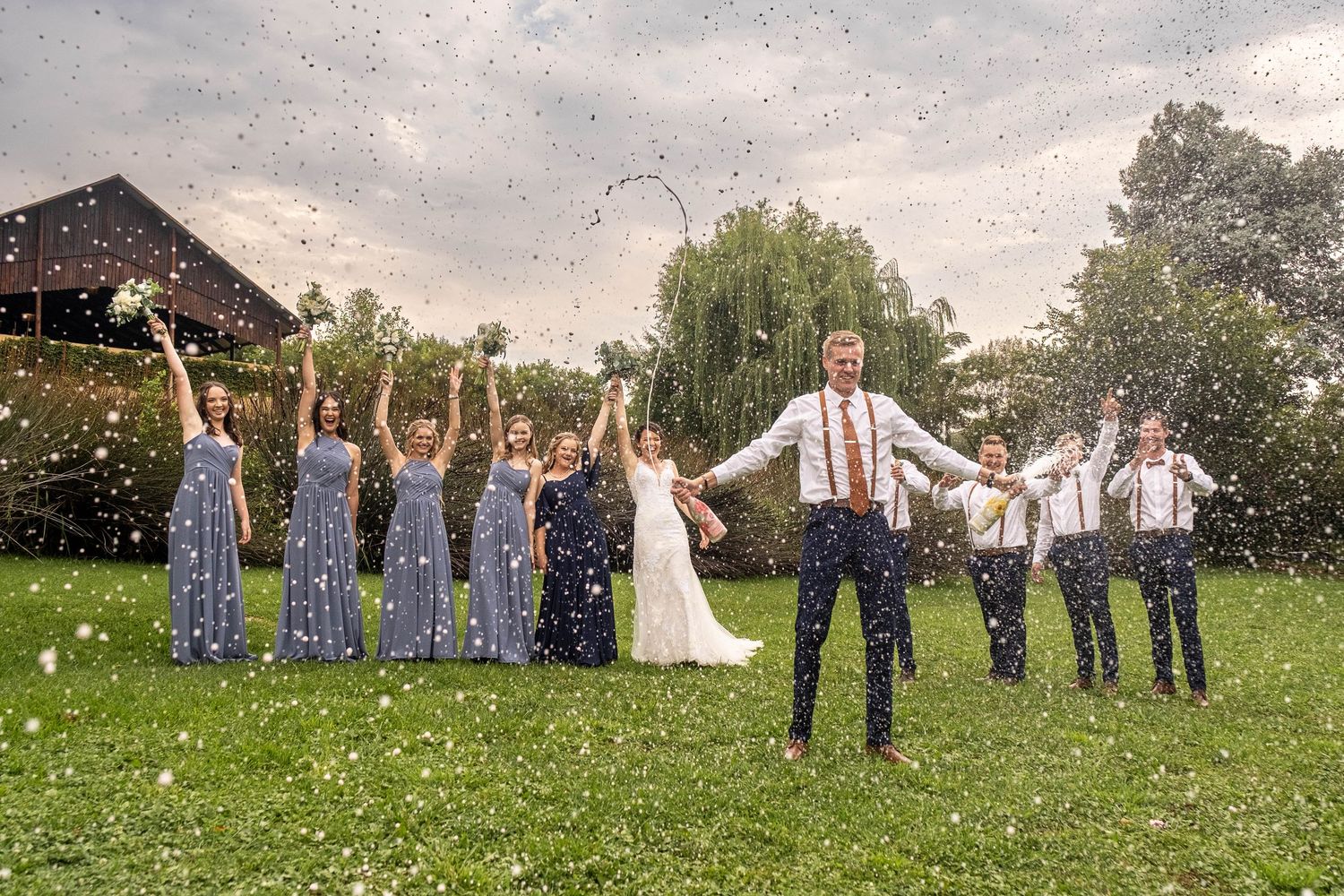 Wedding party celebrates on green lawn with confetti flying as bride and groom stand center surrounded by bridesmaids and groomsmen.
