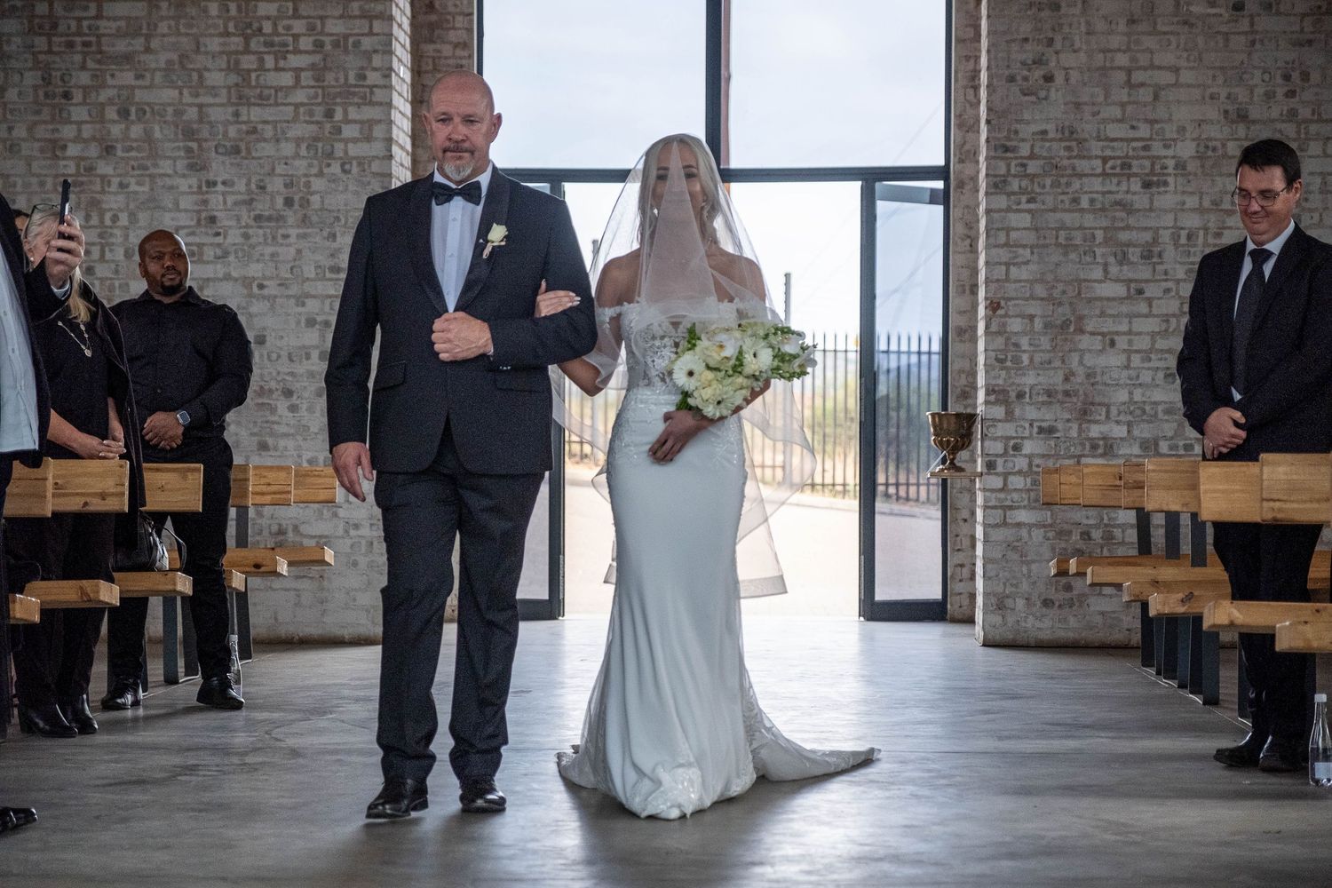 A bride in a white gown walks down the aisle with her father in a modern brick-walled wedding venue.