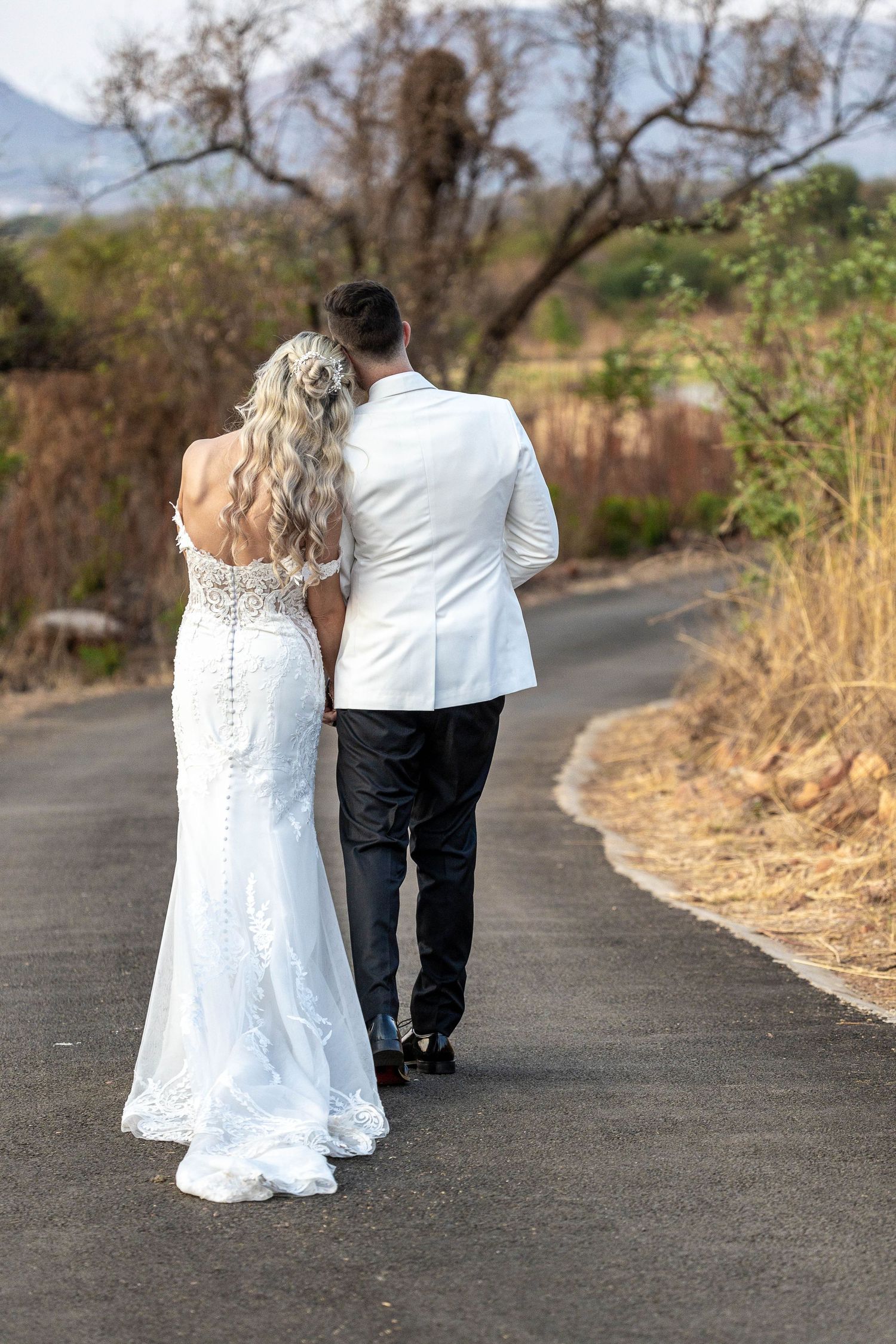 Bride and groom walk away hand-in-hand down a winding road surrounded by dry African bushveld scenery.