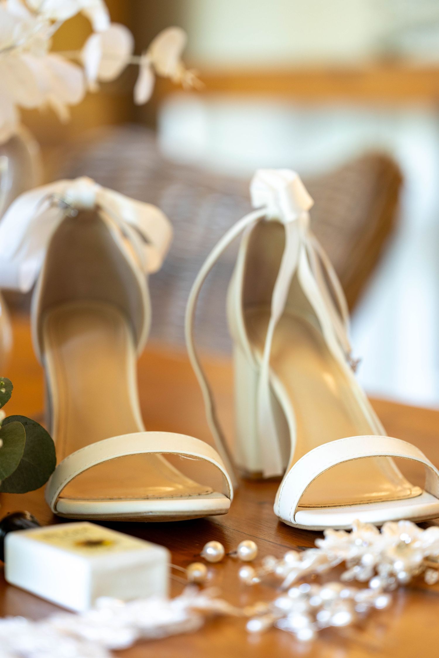 Close-up of elegant white bridal heels with ribbon ties surrounded by pearl jewelry on a wooden surface.