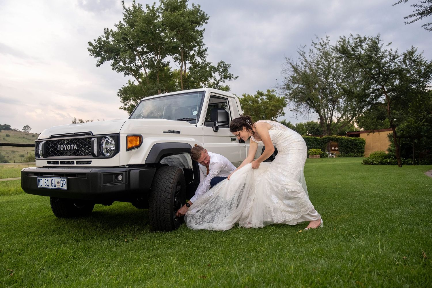 Bride assists groom who playfully lies under a white Toyota Land Cruiser on a green lawn at their wedding.