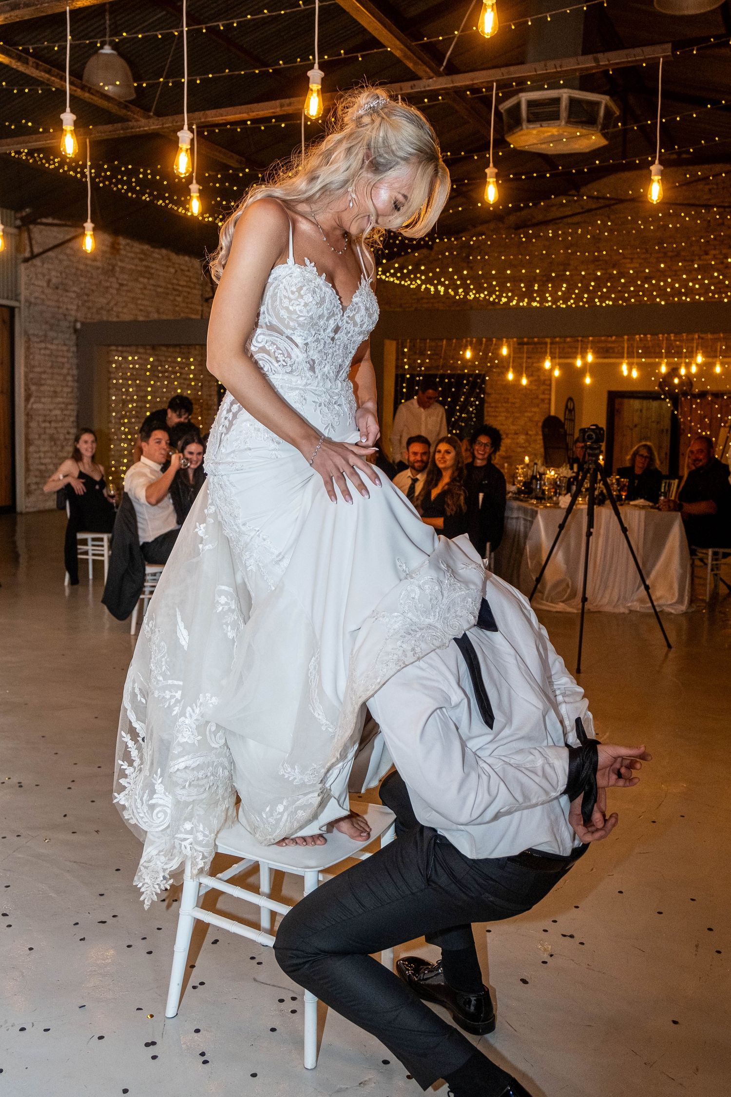 Groom reaching under bride's dress for garter while she stands on white chair at reception.
