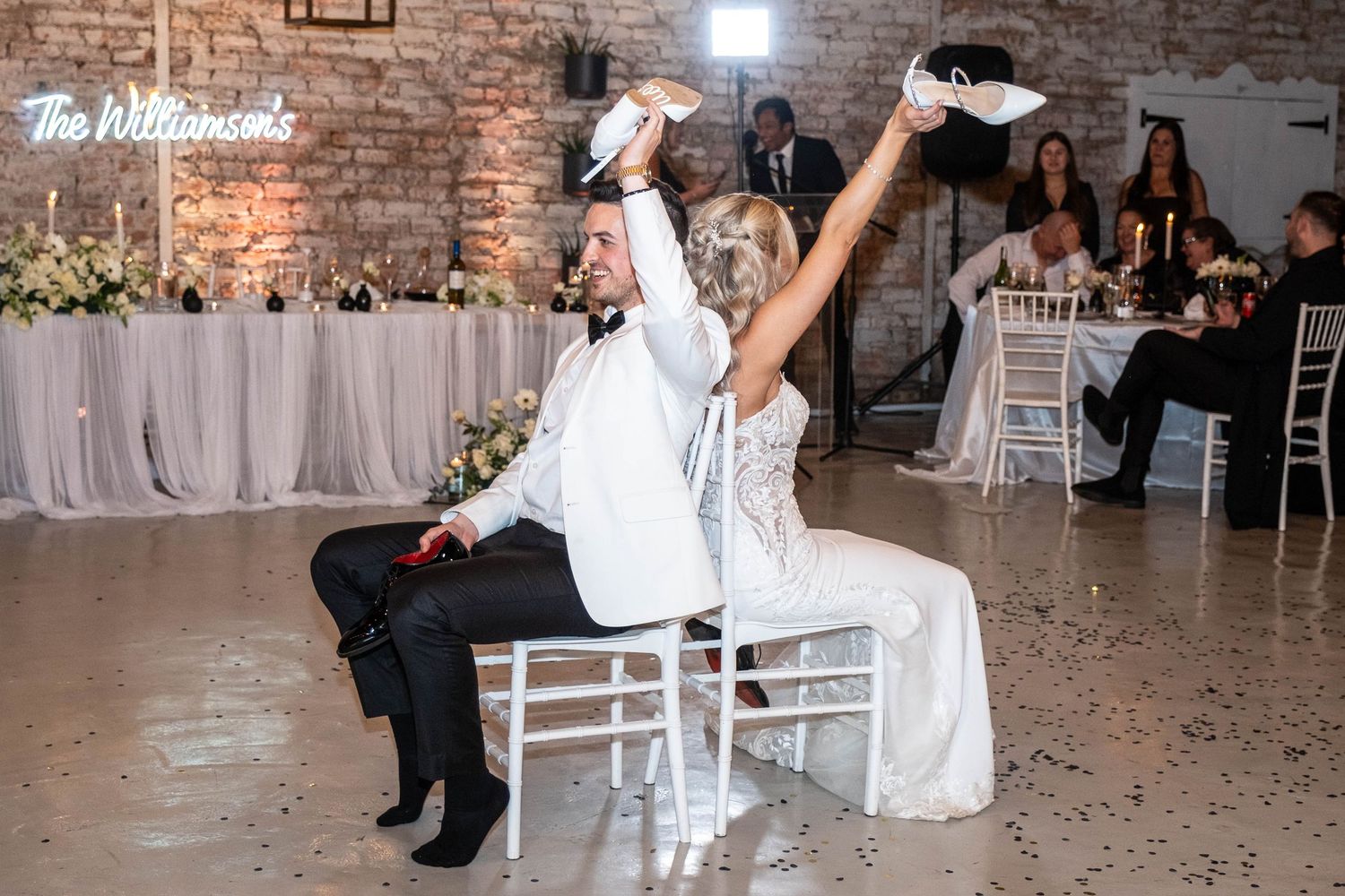Bride and groom play fun wedding shoe game seated in chairs at rustic barn reception.