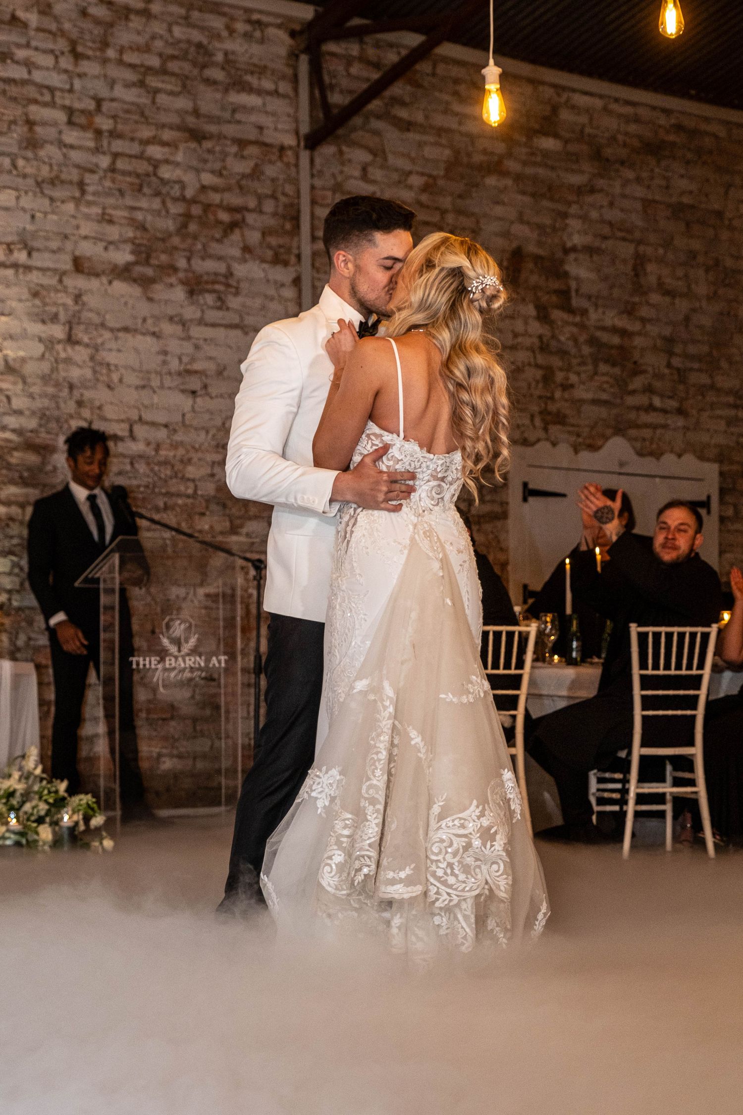 Bride and groom sharing first dance kiss surrounded by dramatic dry ice fog in barn venue.